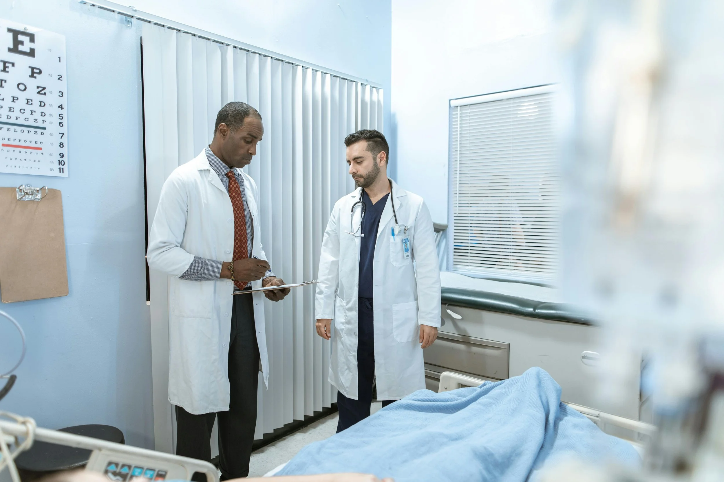 Two doctors in a hospital room, one holding a clipboard and the other with a stethoscope, near a hospital bed with a patient's foot visible under a blue blanket.