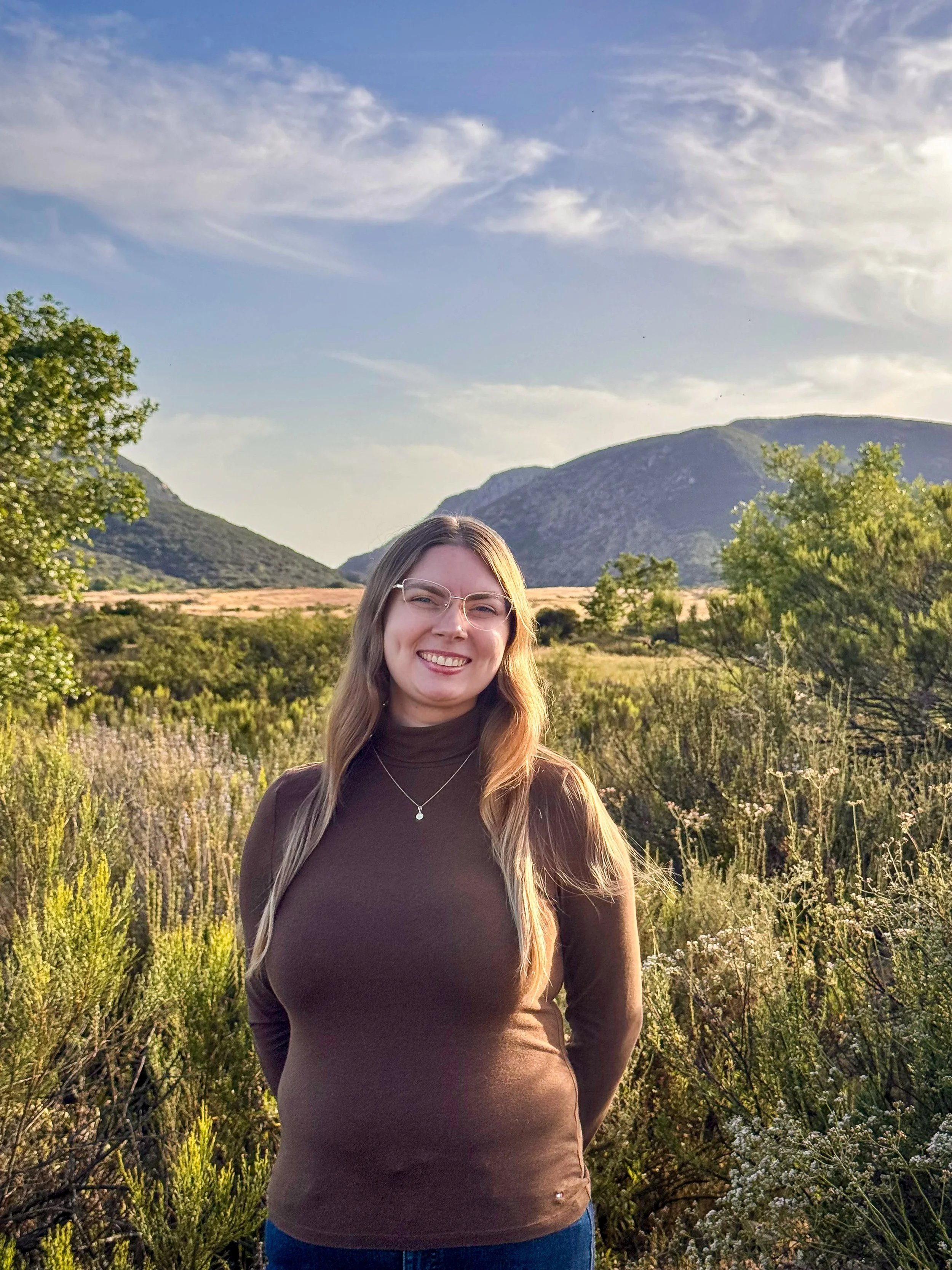 A woman with long blonde hair, glasses, and a brown turtleneck sweater smiling outdoors in a natural landscape with mountains, trees, and bushes under a partly cloudy sky.