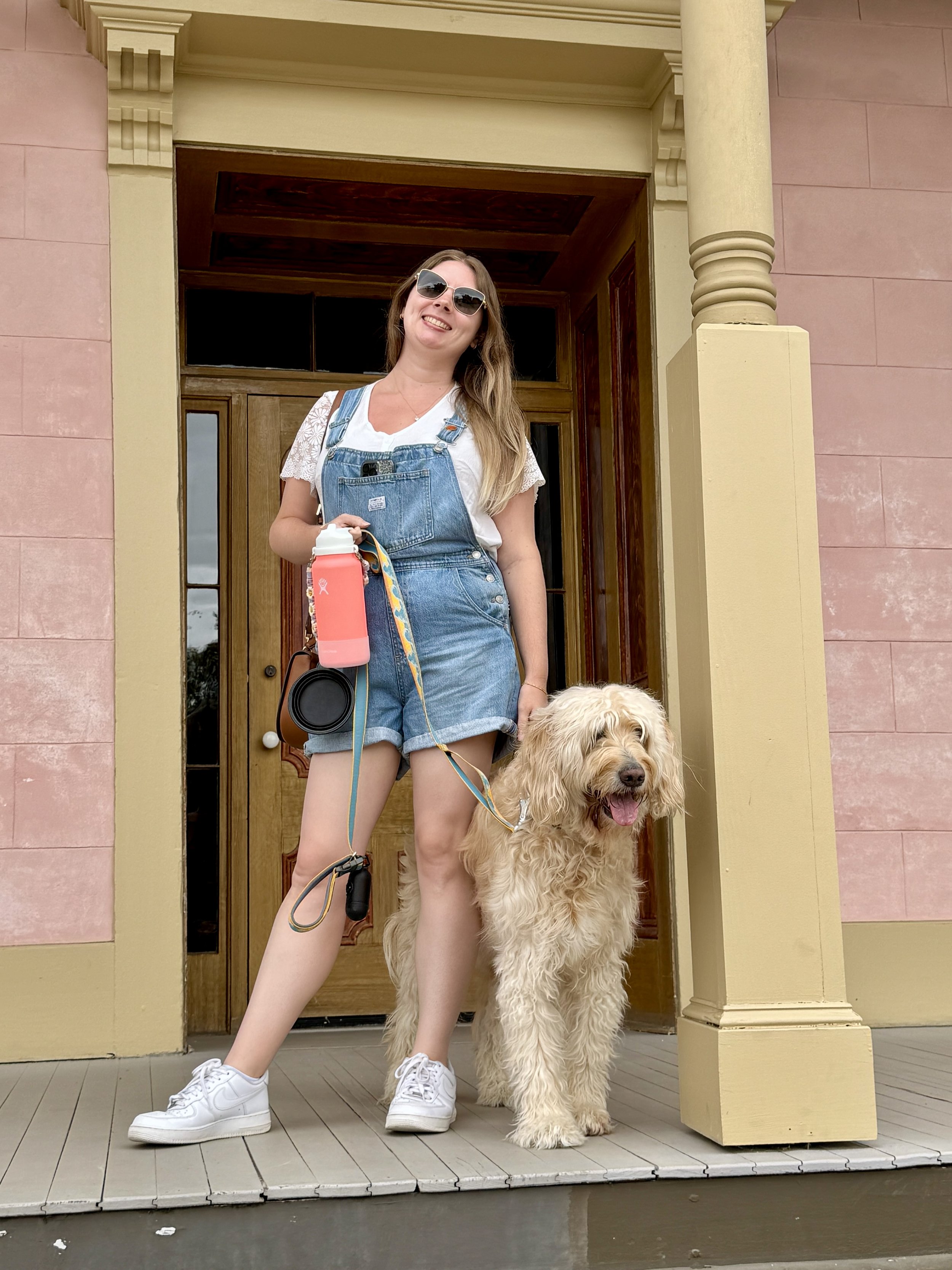 A woman wearing sunglasses and denim overalls standing on a porch with a large, fluffy dog. The woman is smiling, holding a water bottle, and dressed casually in a white t-shirt and sneakers. The dog has light-colored, curly fur and is sitting next to her.