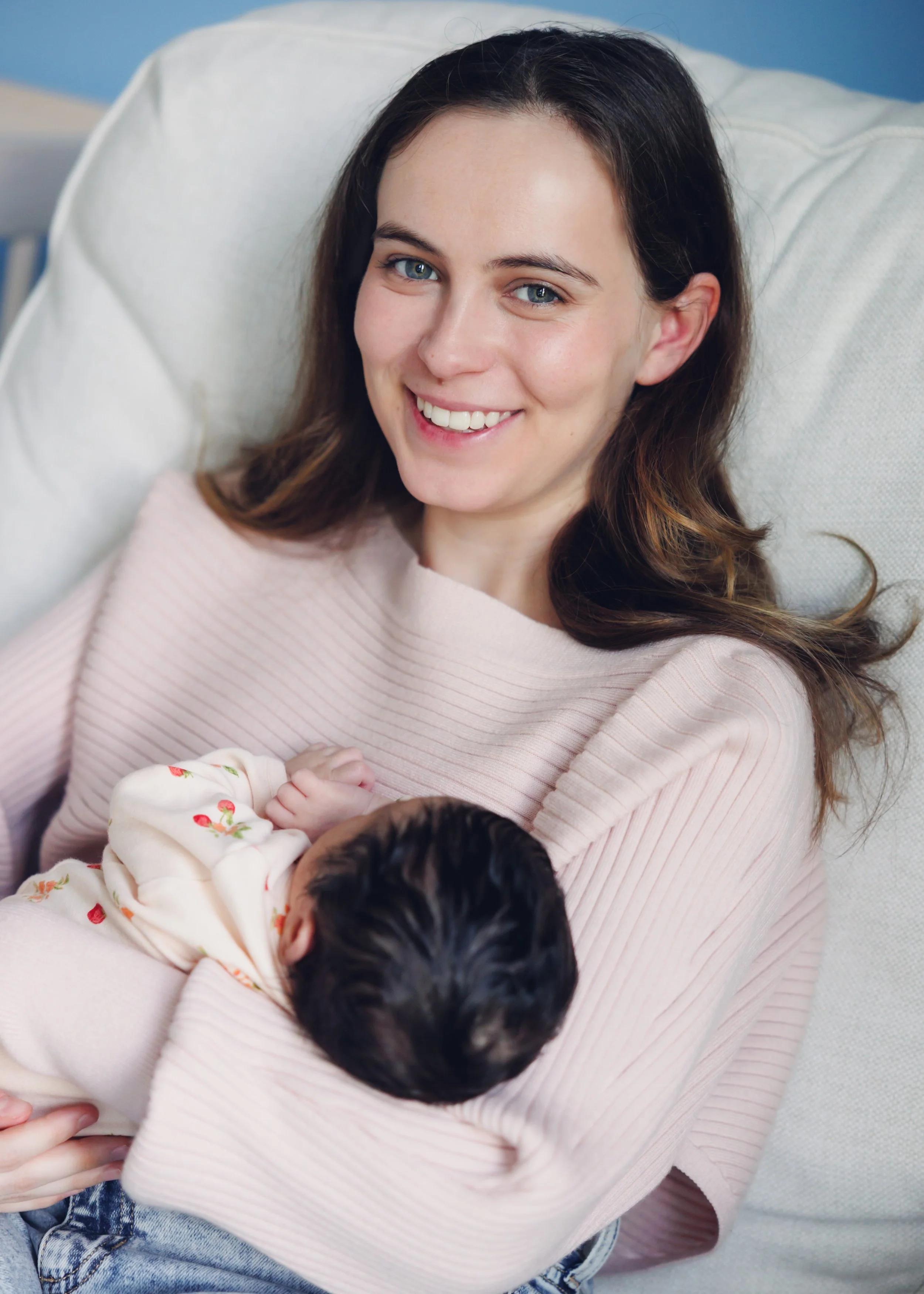 A young woman with long brown hair and blue eyes smiling while holding a newborn baby in her arms, sitting on a light-colored sofa.