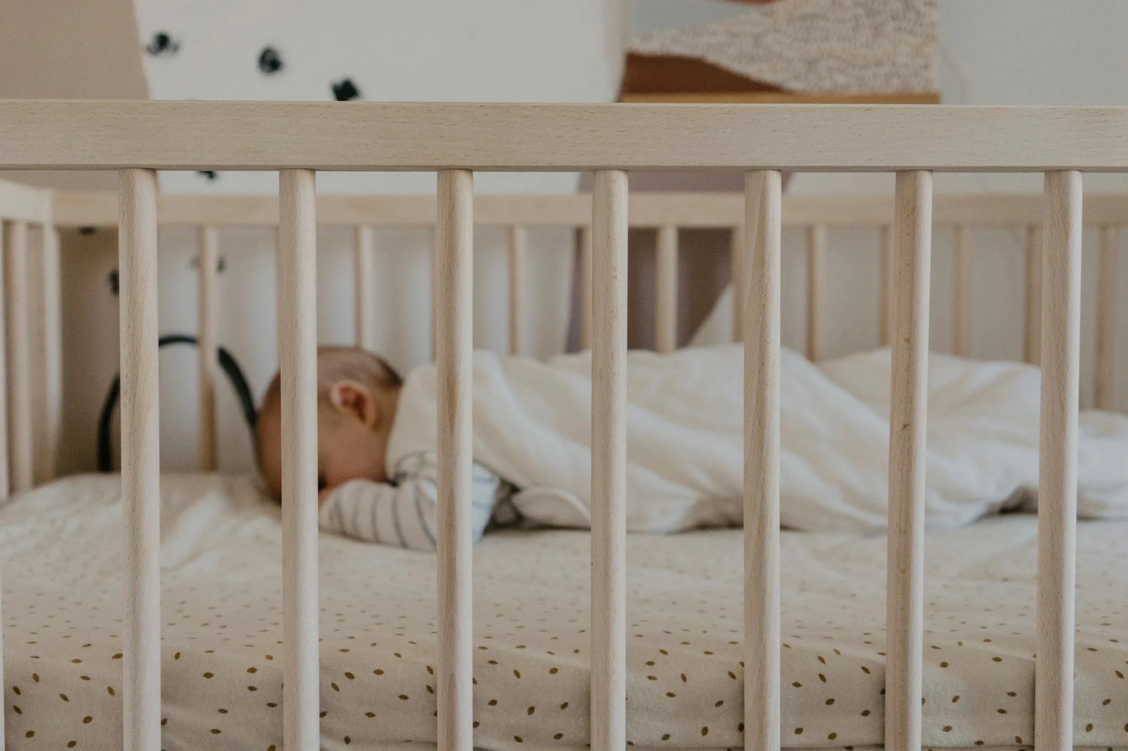 A baby sleeping in a crib with wooden bars, lying on a dotted sheet with a pillow.