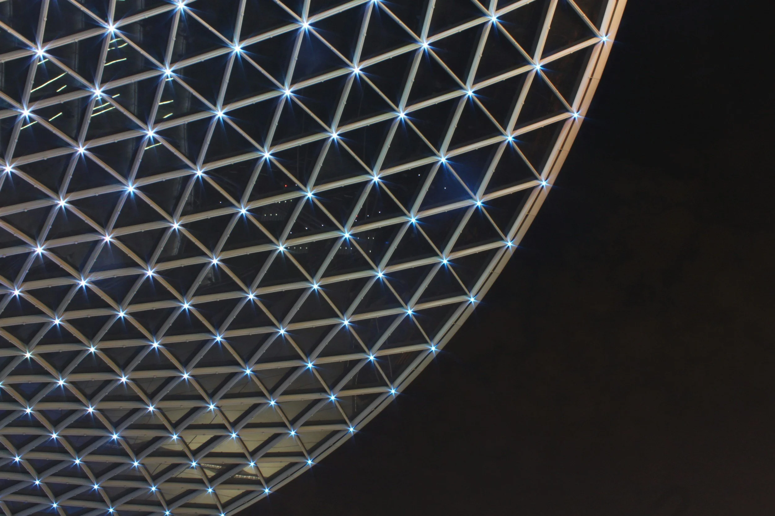 Night view of a modern building with a geometric lattice pattern illuminated by blue and white lights, against a dark sky.