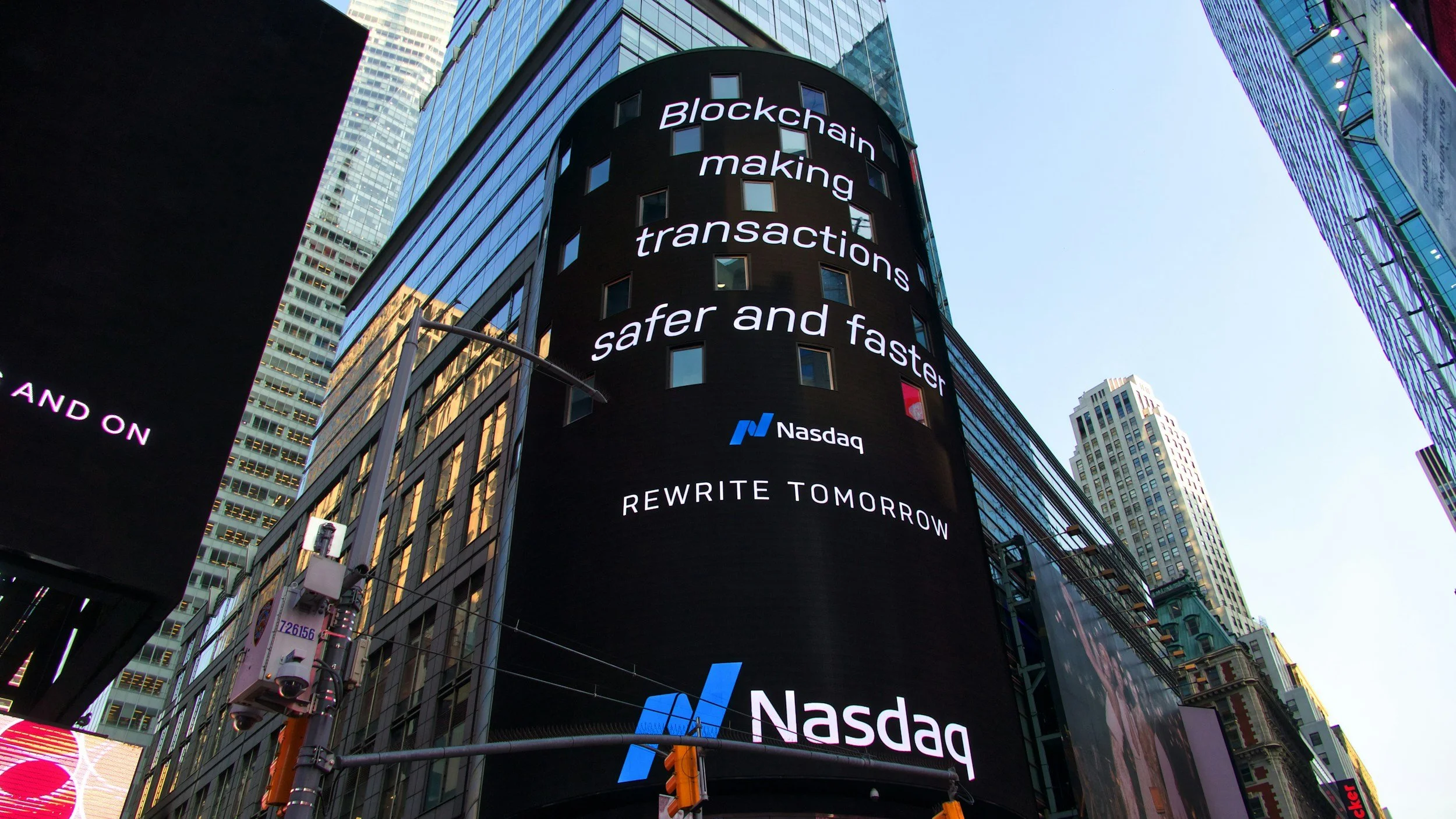 A large digital billboard displays an advertisement on the side of a tall building in Times Square, New York City. The text on the billboard reads: 'Blockchain making transactions safer and faster,' with the Nasdaq logo and the slogan 'Rewrite Tomorrow' underneath.