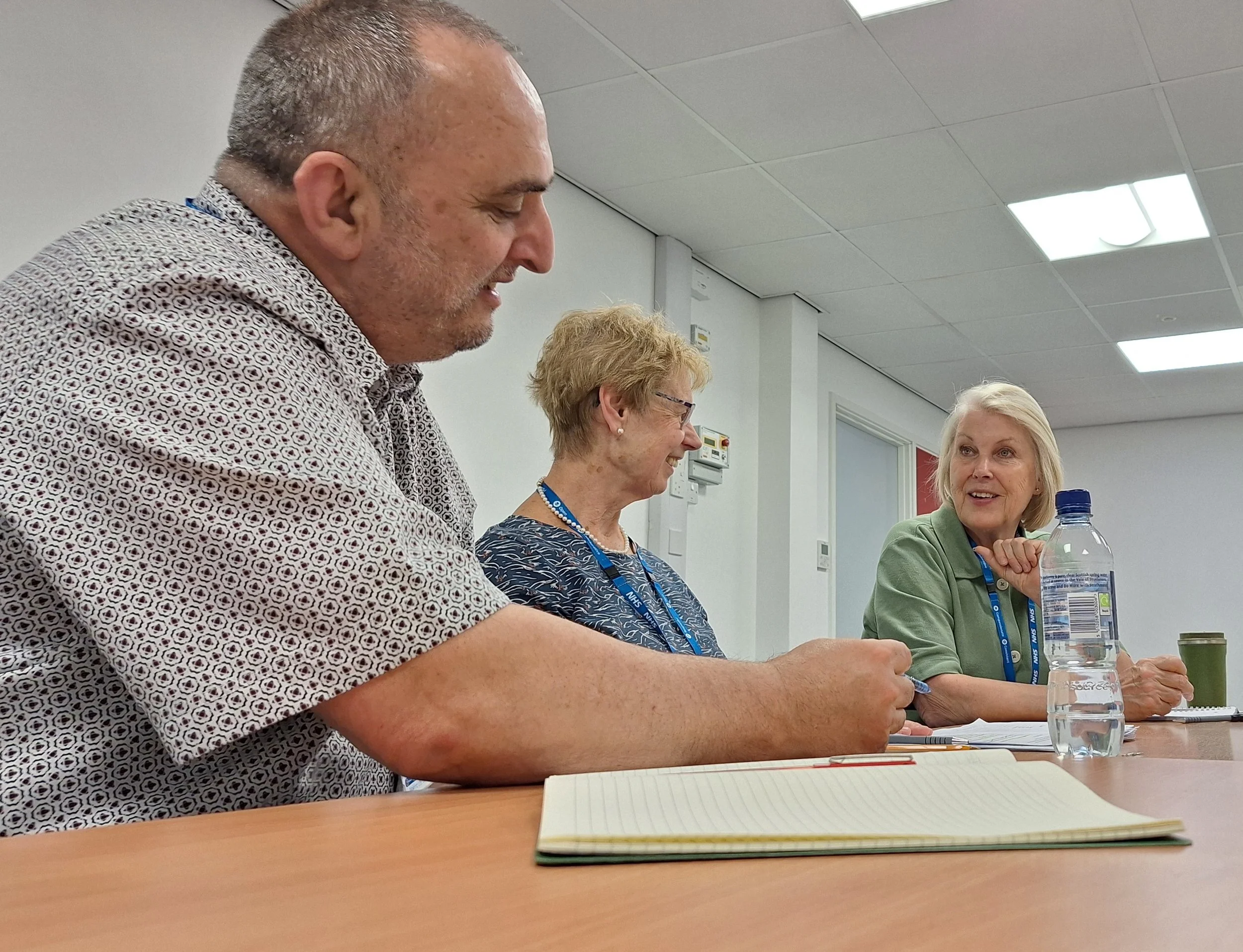 Three older adults sitting at a conference table engaged in conversation, with notebooks, pens, a water bottle, and a coffee mug on the table.