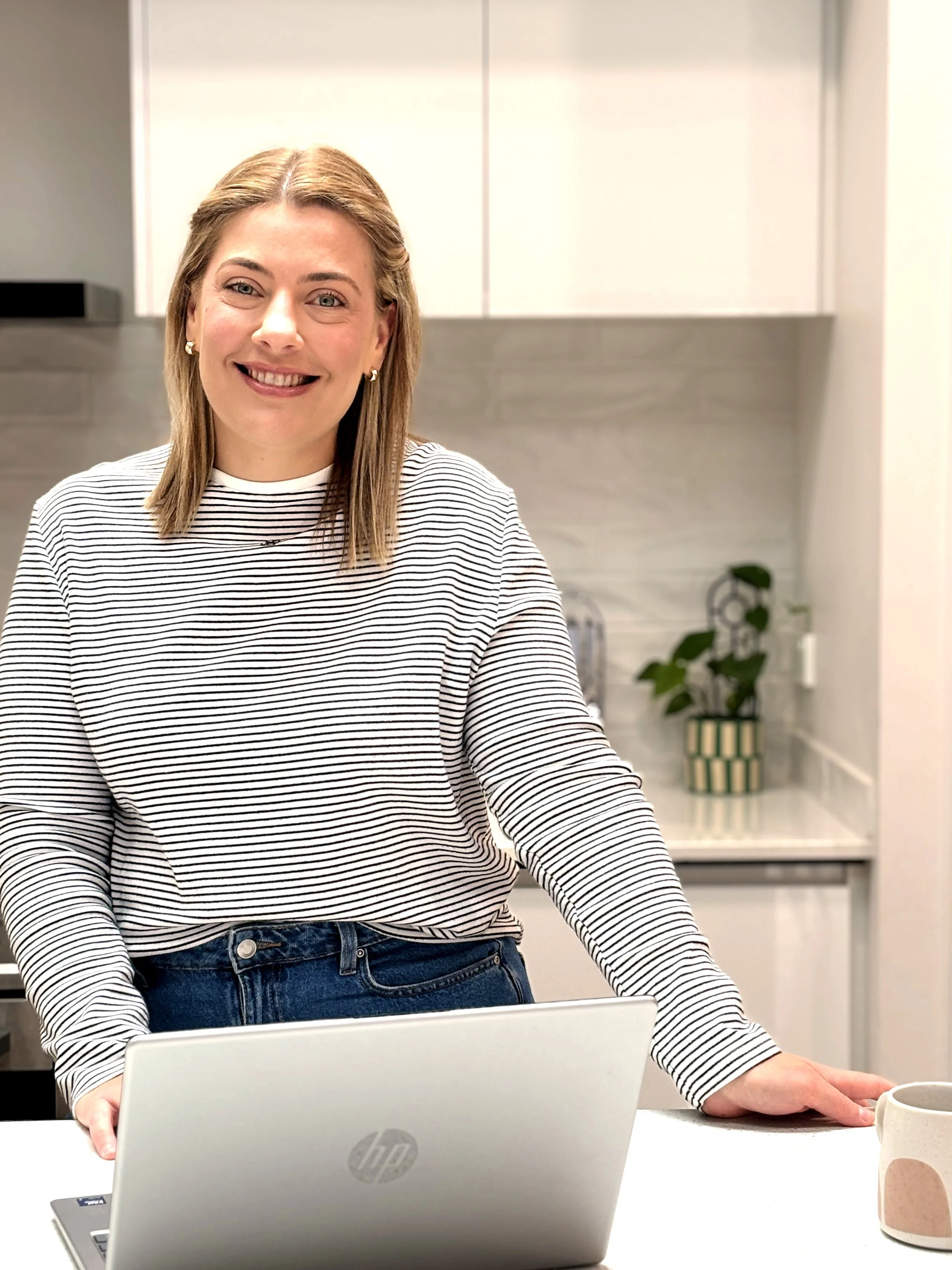Smiling woman with blonde hair in a striped shirt sits at a table with her hand on a white laptop in a modern kitchen, with cabinets and a potted plant in the background.