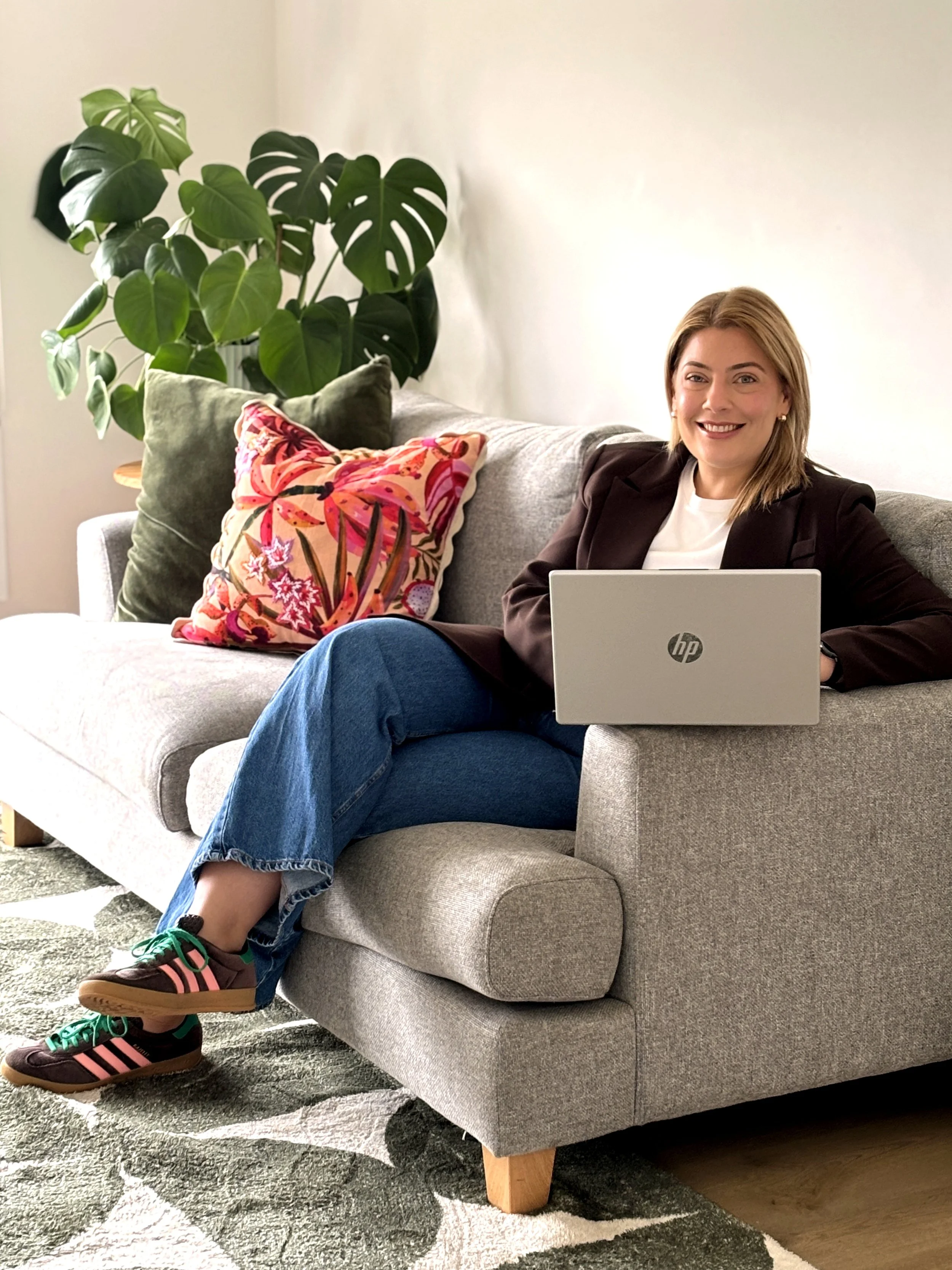 A woman sitting on a light gray sofa, smiling, with a silver HP laptop on her lap. She is wearing a white shirt, a dark blazer, blue jeans, and colorful sneakers. There are decorative pillows on the sofa, one with a pink and orange floral pattern, another that is plain green. Behind her, a large green monstera plant is on the left side of the image, and the background consists of a plain, light-colored wall. The sofa is on a textured gray and white rug.