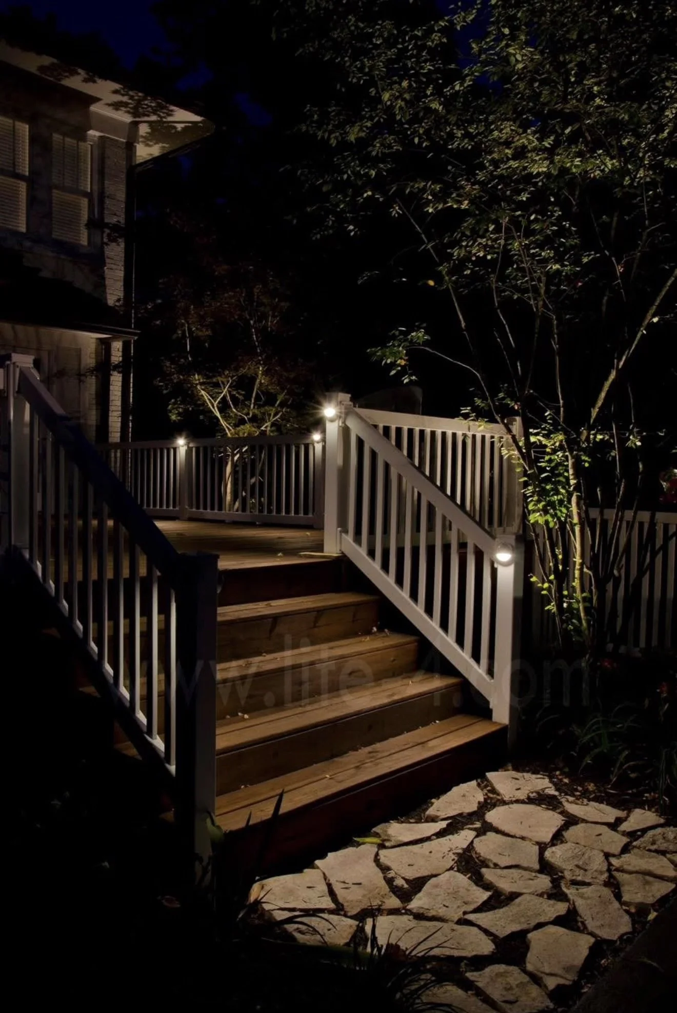 Night view of a wooden stairs with white railings, illuminated by small lights, leading to a deck area in a garden with trees and a stone pathway.