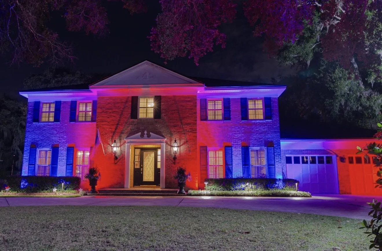 A large two-story house illuminated with red, white, and blue lights at night, with a well-maintained lawn, bushes, and trees surrounding it.