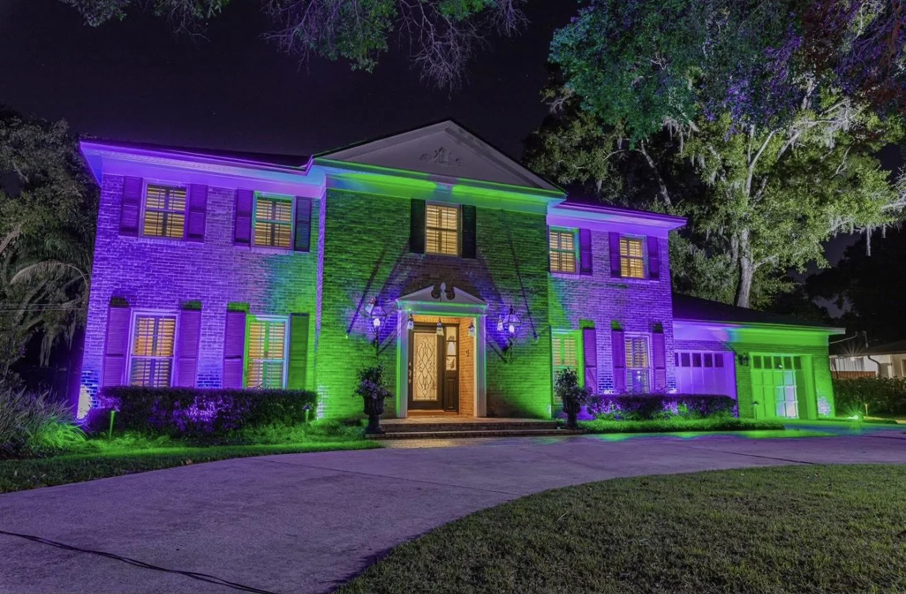 A house illuminated with colorful purple, green, and blue lights at night, with trees in the background and a curved driveway in the foreground.