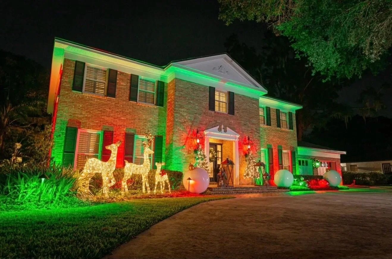 A large brick house decorated with Christmas lights in red, green, and white, including reindeer figures, illuminated trees, and oversized ornaments, with a dark sky background.