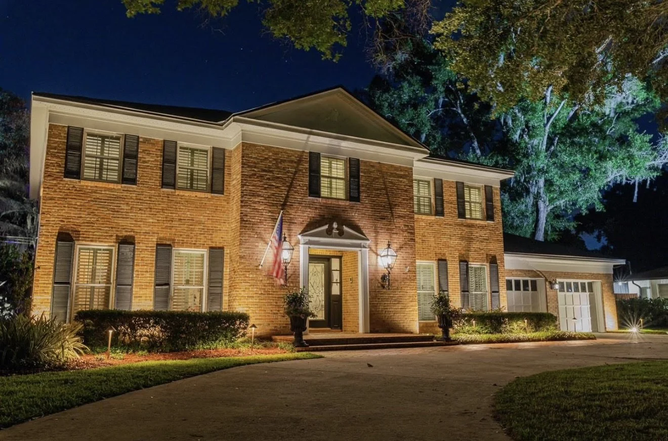 A well-lit brick house at night with multiple windows, shutters, and a front door with a lantern light. There are small garden bushes, potted plants, and outdoor lighting illuminating the landscaping, with a driveway leading to an attached garage. A large tree is visible in the background under a dark, starry sky.