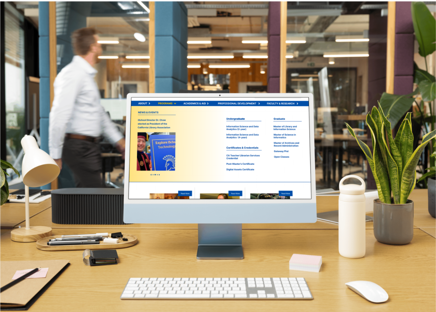 Computer on a desk displaying a college website with program and course information, in a modern office with plants and a blurred person in the background.