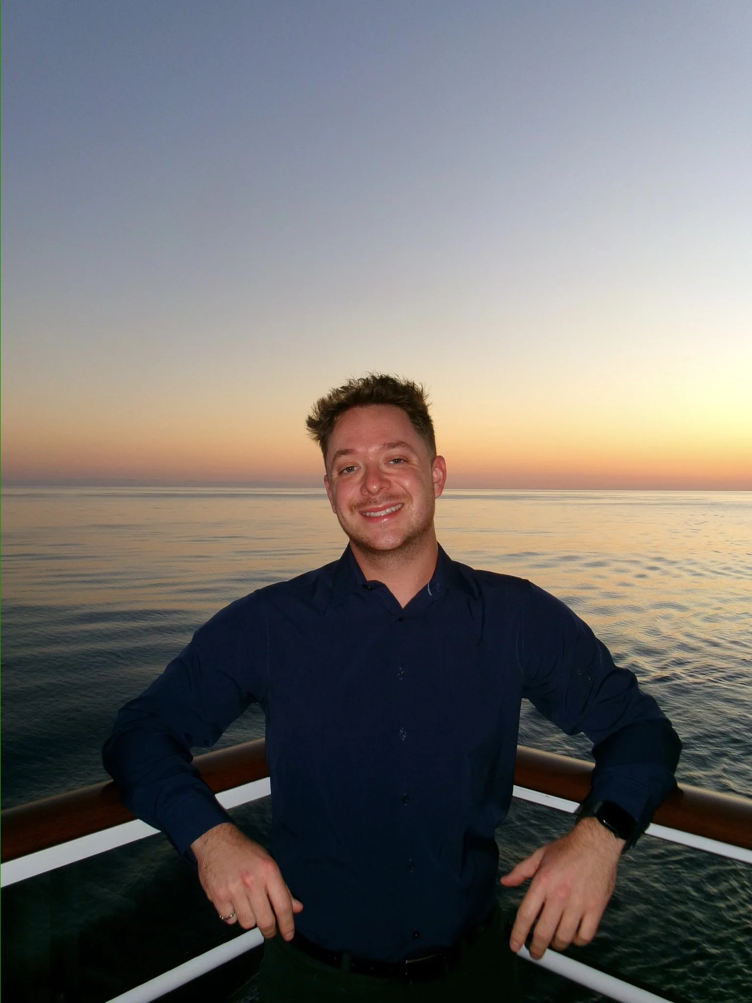 A young man with light skin, brown hair, and a short beard, smiling and standing on a boat railing with his hands resting on it, during sunset over calm ocean water.