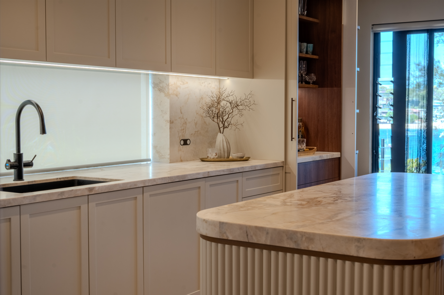 Modern kitchen with beige cabinets, marble countertops, and a window with blue curtains. There is a black faucet, decorative vases, and a tree branch on the counter.