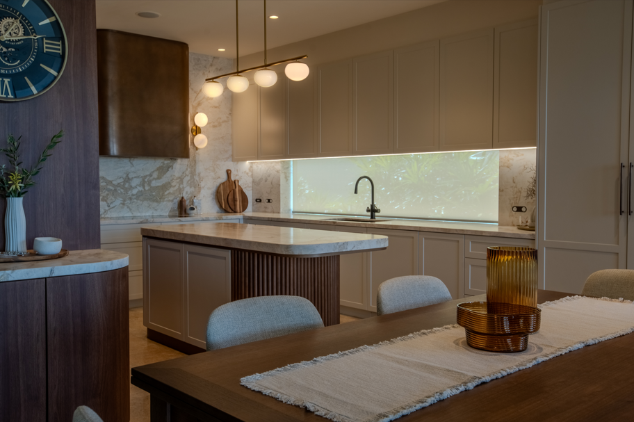 Modern kitchen featuring beige cabinets, a marble backsplash, a central island, pendant lighting, a window with greenery outside, and a dining table with woven vases.