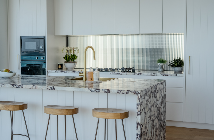 Modern kitchen with white cabinets and marble accents, featuring a gold faucet, potted plants, and a breakfast bar with three wooden stools.