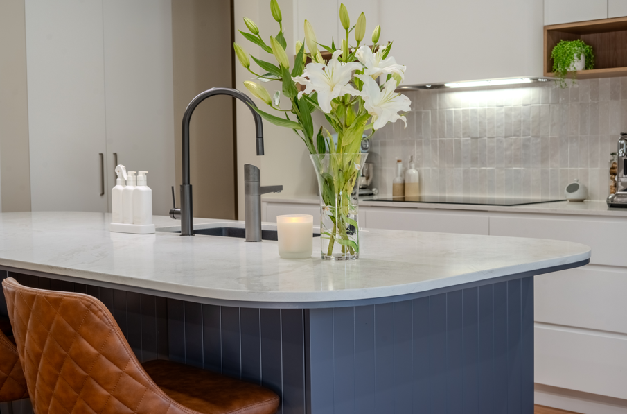 Kitchen with a large white marble island, a black faucet, a glass vase with white lilies, a lit candle, and a brown quilted leather chair.