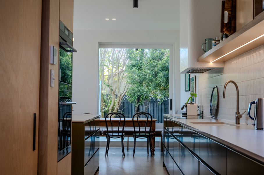 Modern kitchen with a large window, black chairs at a wooden table, white countertops, a stainless steel faucet, and open shelving with kitchen utensils.