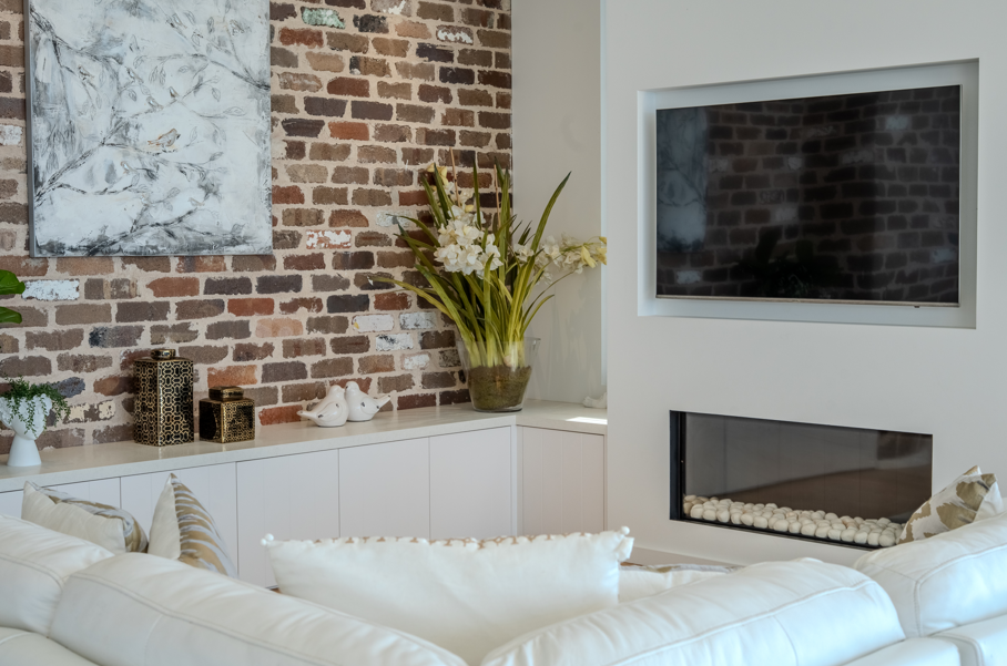 Living room with a white couch, decorative pillows, a large bouquet of white flowers, a sculptural piece, and a brick wall behind a white built-in cabinet. A flat-screen TV is mounted on a white wall with a modern fireplace below it.