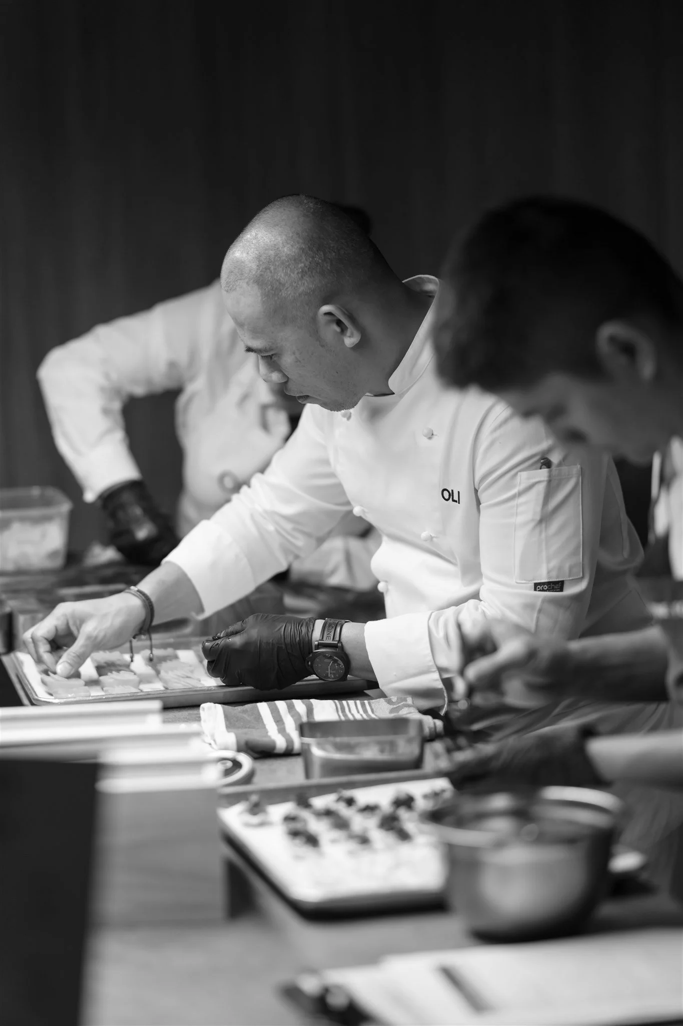 Black and white photo of chefs in a kitchen, focused on preparing food, with one chef wearing gloves and looking down at ingredients or food on a tray.