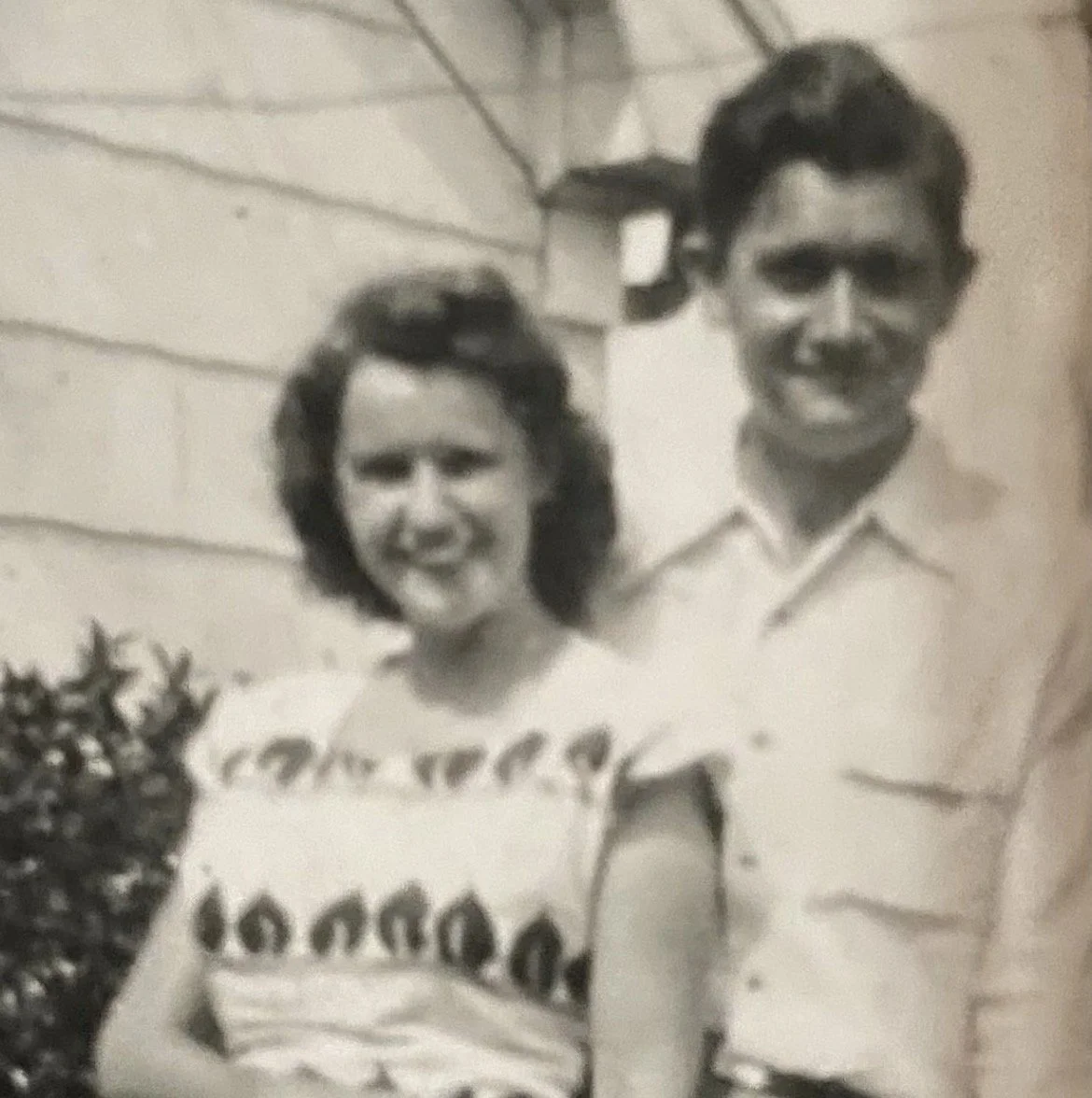 A black and white photograph of a woman and a man standing outside in front of a wooden house. The woman is smiling and wearing a sleeveless top with a pattern, while the man is also smiling, wearing a collared shirt.