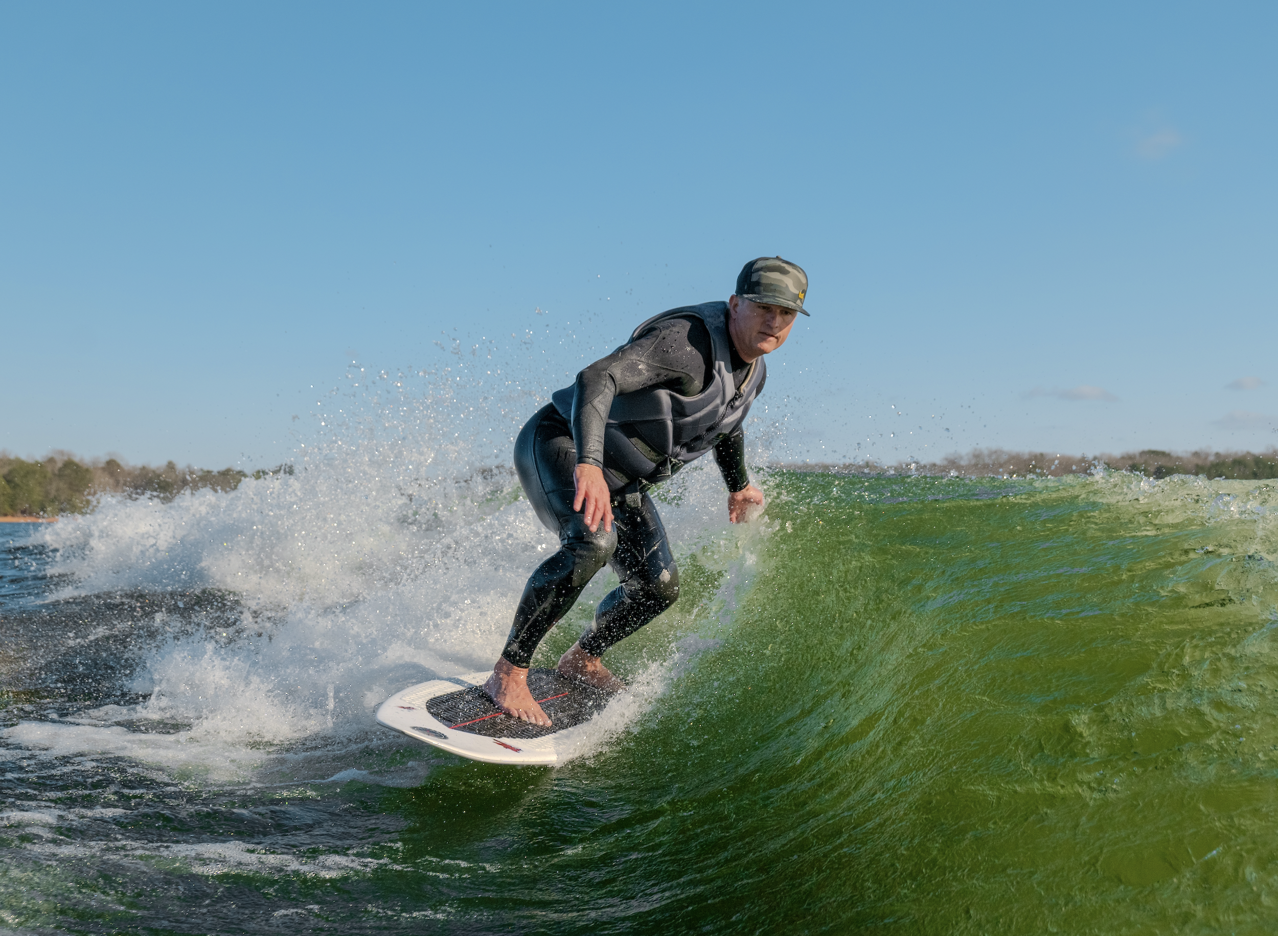 Man surfing on a wave, wearing a wetsuit and cap, with clear blue sky in the background.