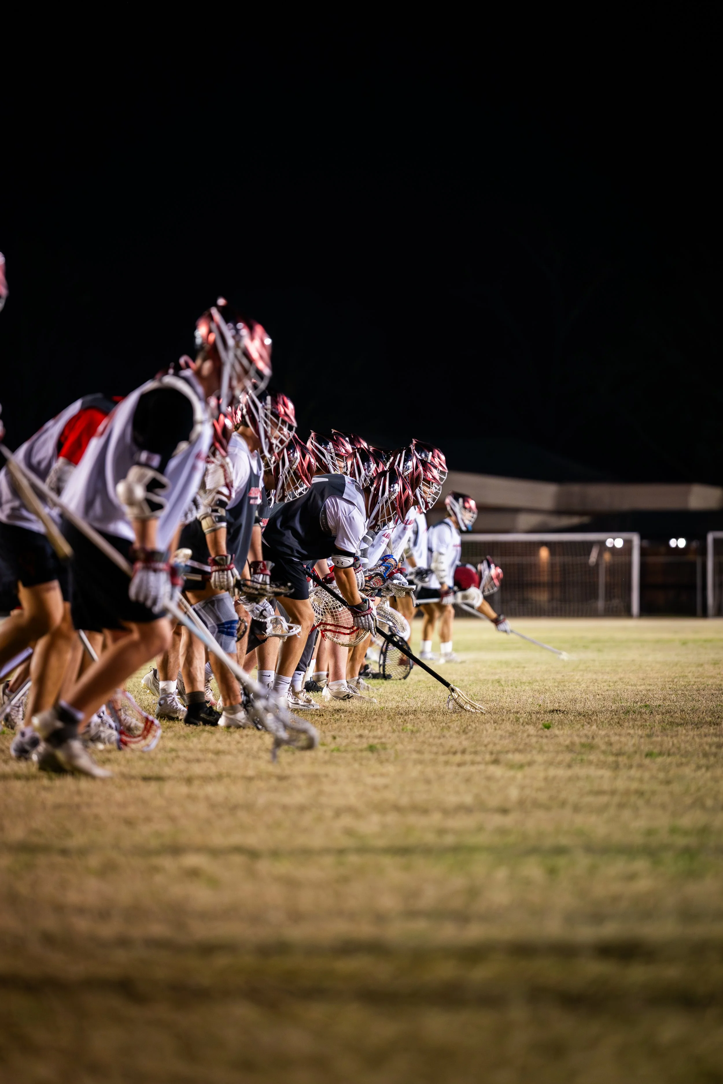 Lacrosse players in uniforms and helmets huddled on the field at night, preparing for a game.