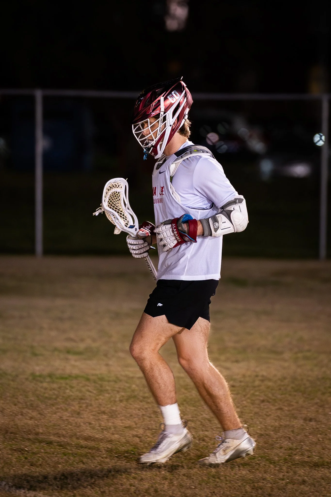 Lacrosse player wearing a helmet, gloves, arm pad, and sports jersey, holding a lacrosse stick, walking on the field during evening or night game.