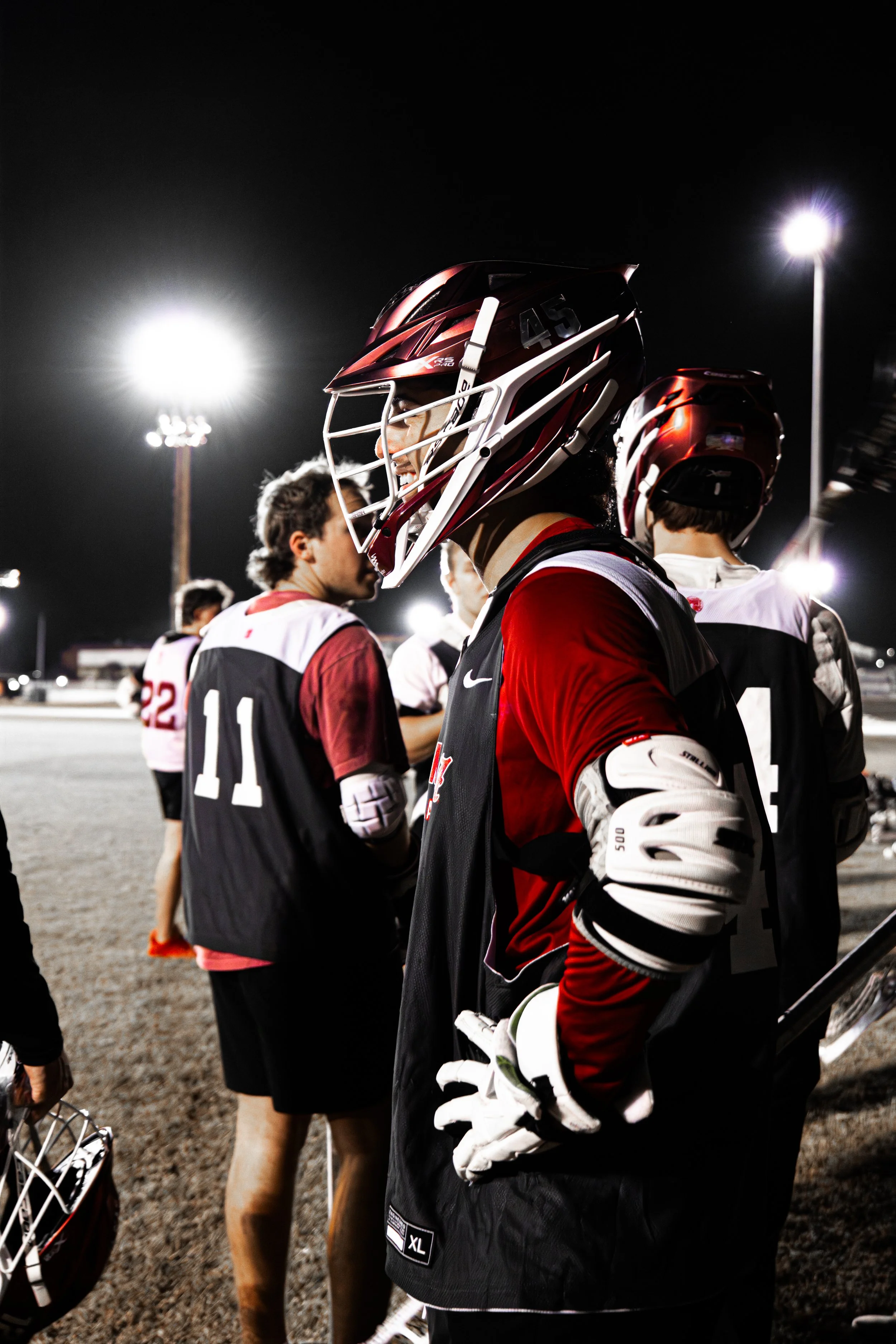 Lacrosse players in gear on a field at night, illuminated by bright stadium lights.