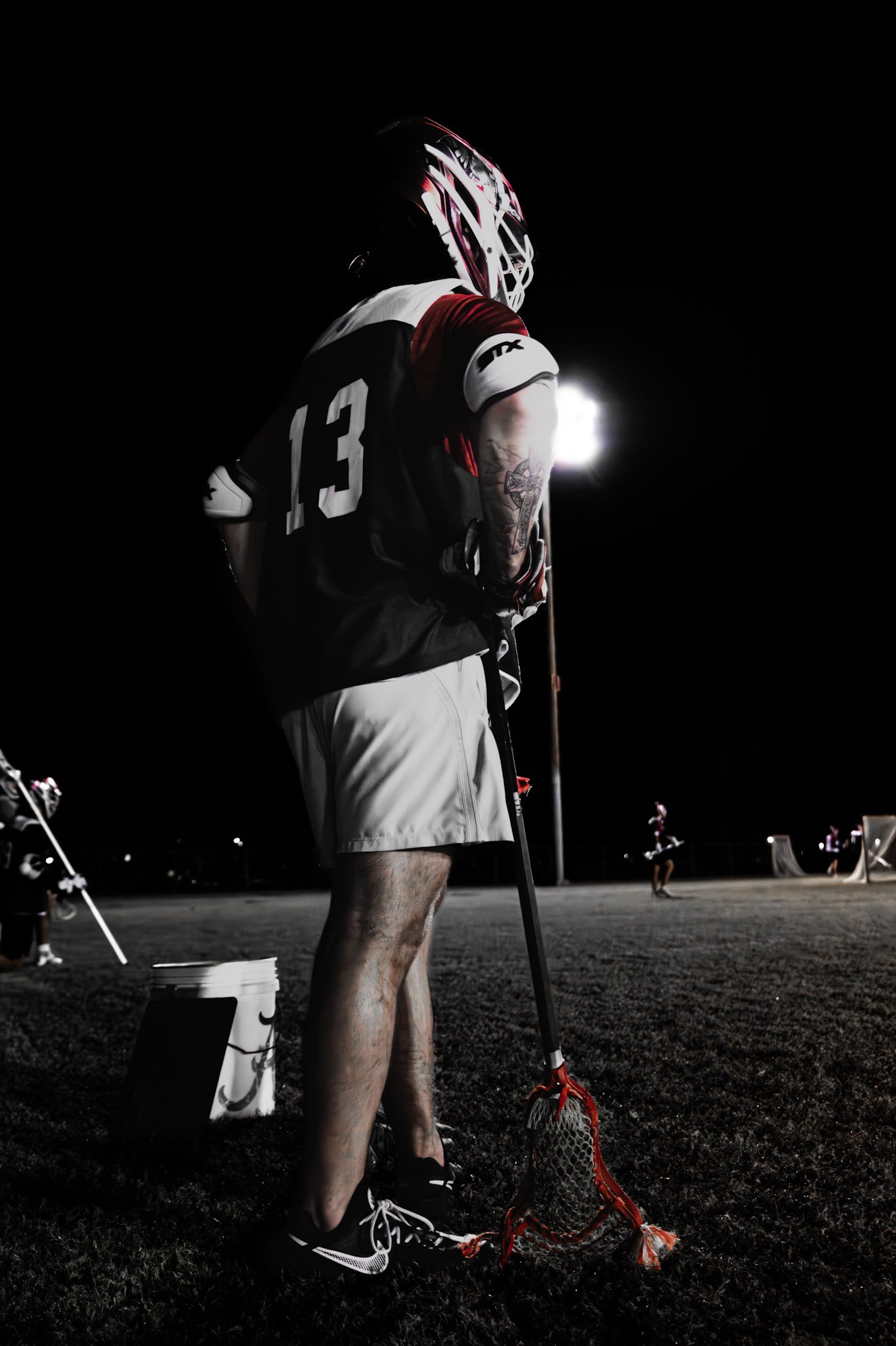 Lacrosse player standing on field at night, wearing a helmet and jersey with the number 13, holding a lacrosse stick with a red net.