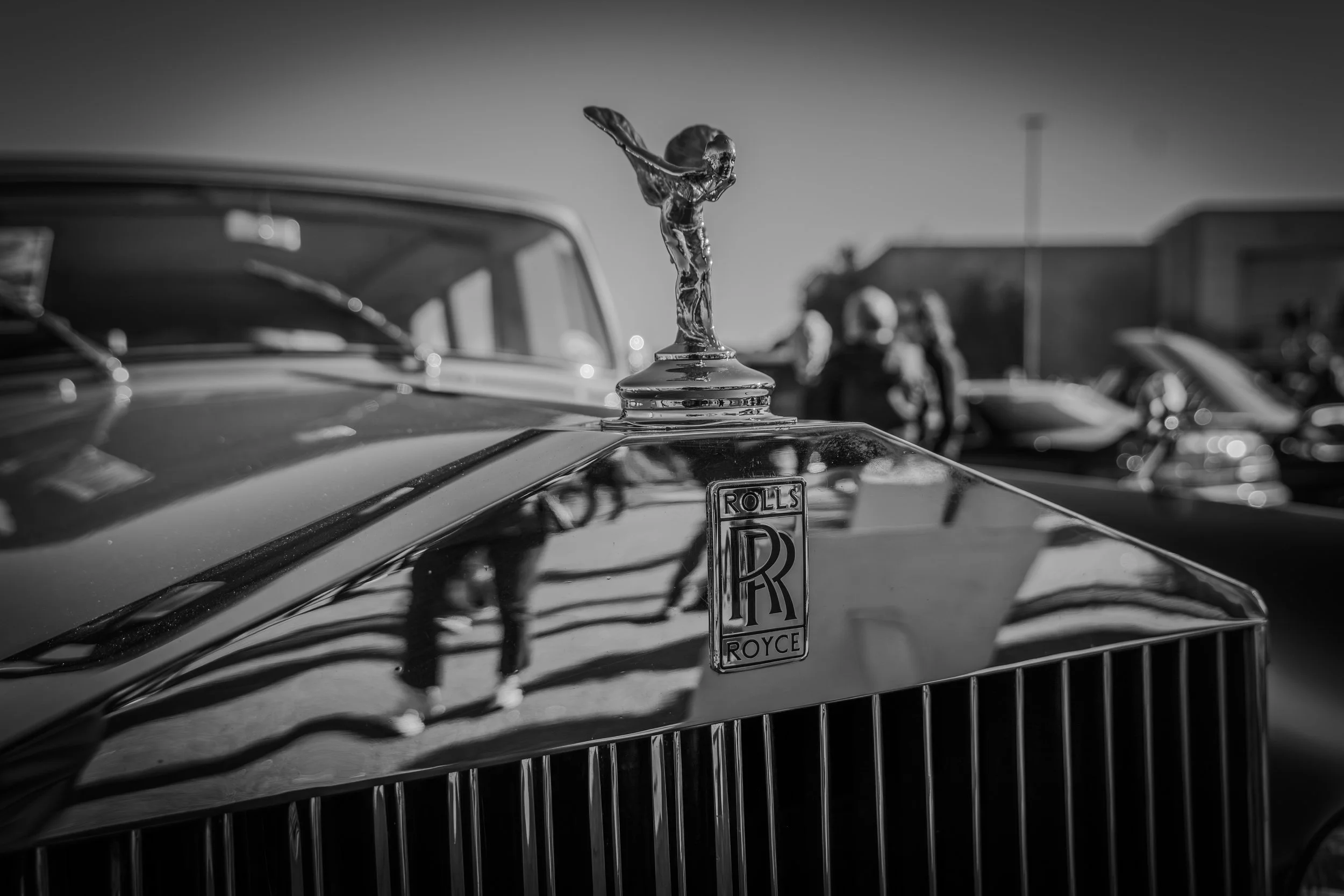 Close-up of the hood ornament on a classic Rolls-Royce car, featuring the Spirit of Ecstasy figure, in black and white with several vintage cars and people in the background.