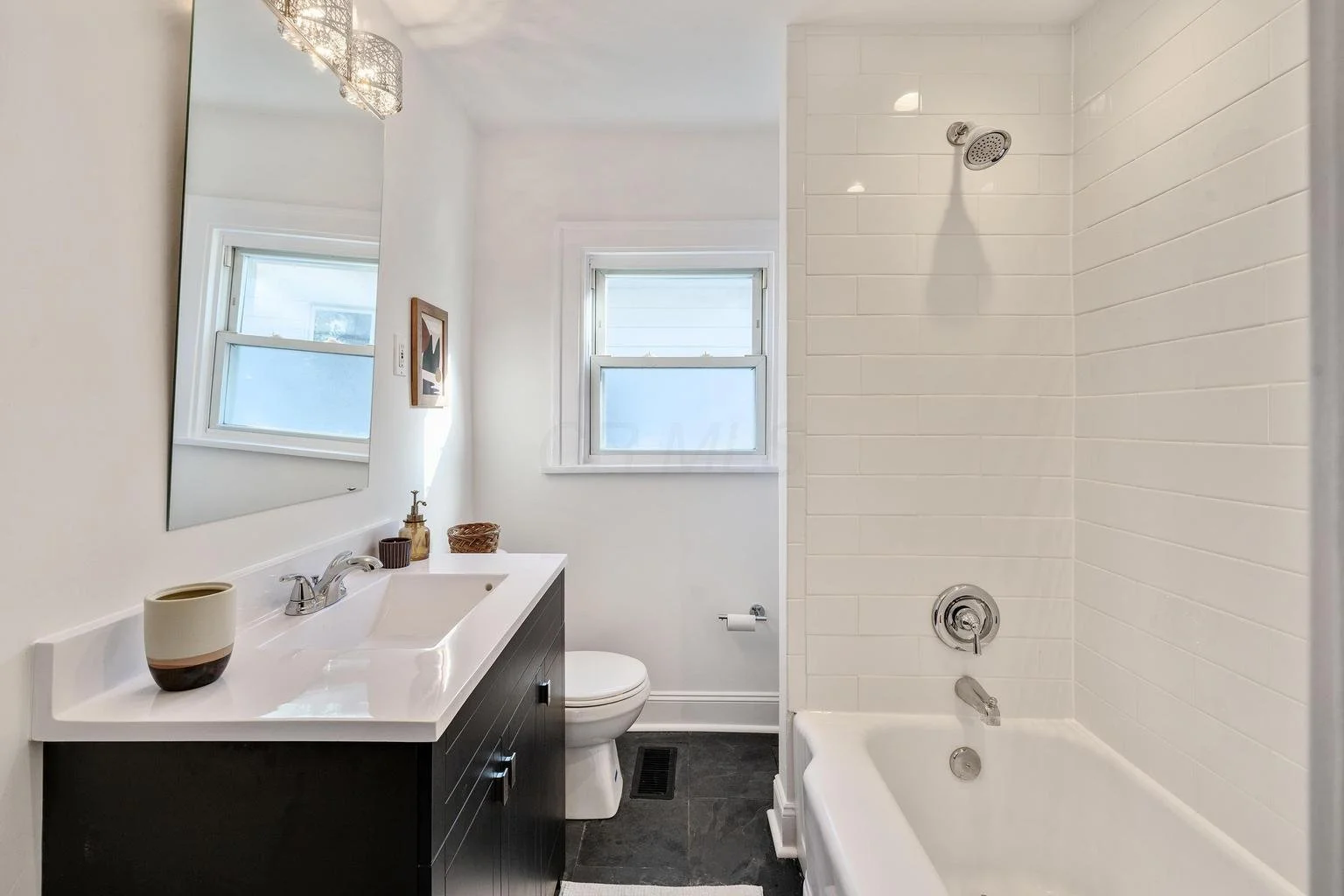 Modern bathroom with a white bathtub and tiled wall, a white toilet, a black vanity with a white sink, a large mirror, two small windows, and decorative items on the vanity.
