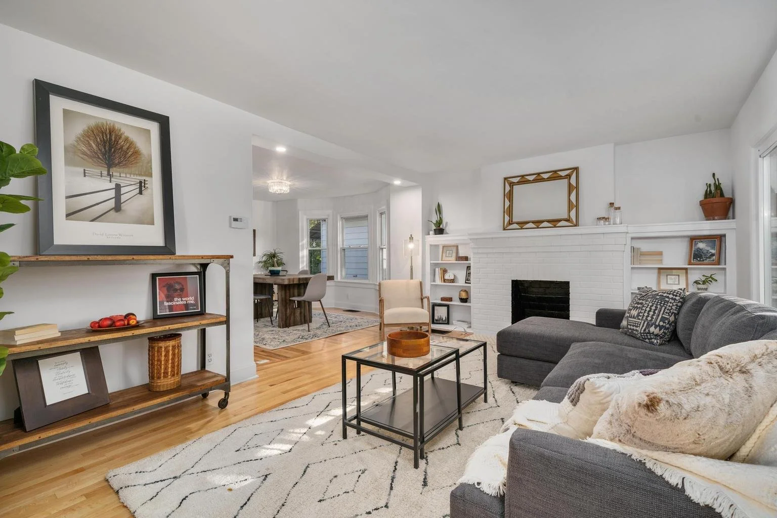 Living room with white brick fireplace, gray sectional sofa, beige armchair, black metal coffee table, white patterned rug, and built-in white bookcases, bright natural light from windows.