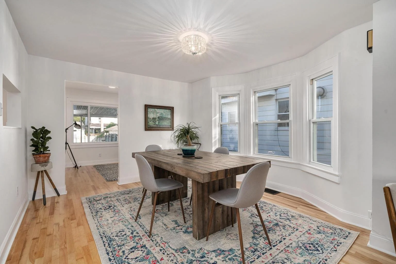 A bright dining room with a wooden table surrounded by four light gray chairs, a potted plant on top, large windows allowing natural light, and a ceiling light fixture casting a starburst pattern.