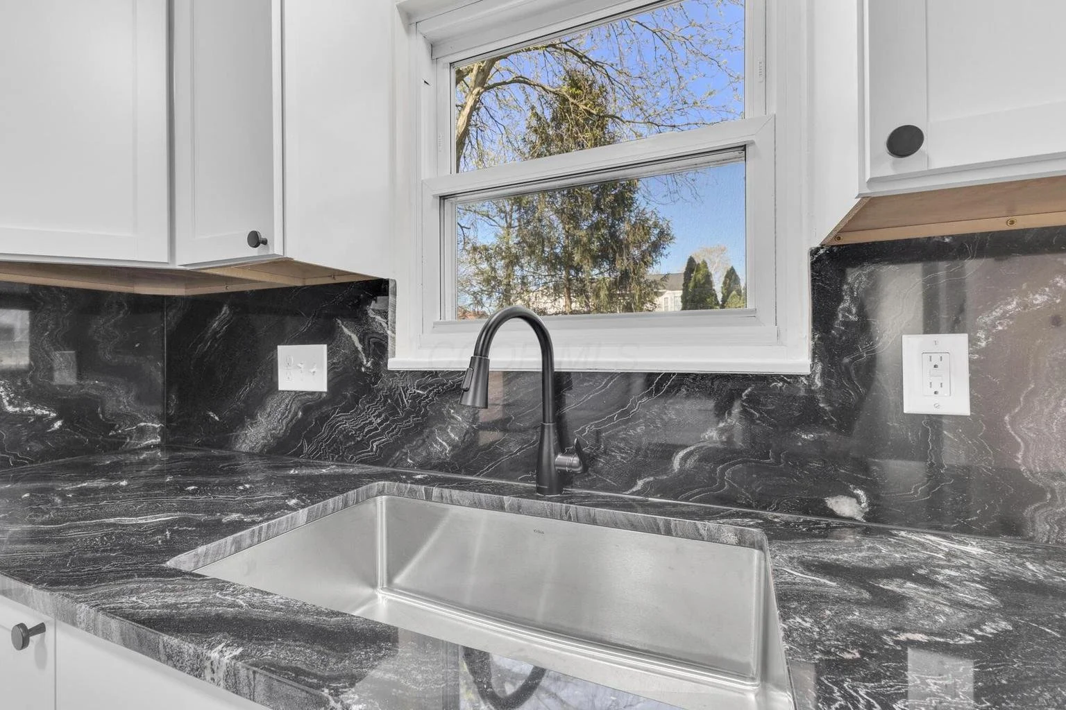 Kitchen sink with a black marble countertop, a black faucet, white cabinets, a double-paned window showing trees outside, and electrical outlets on the backsplash.