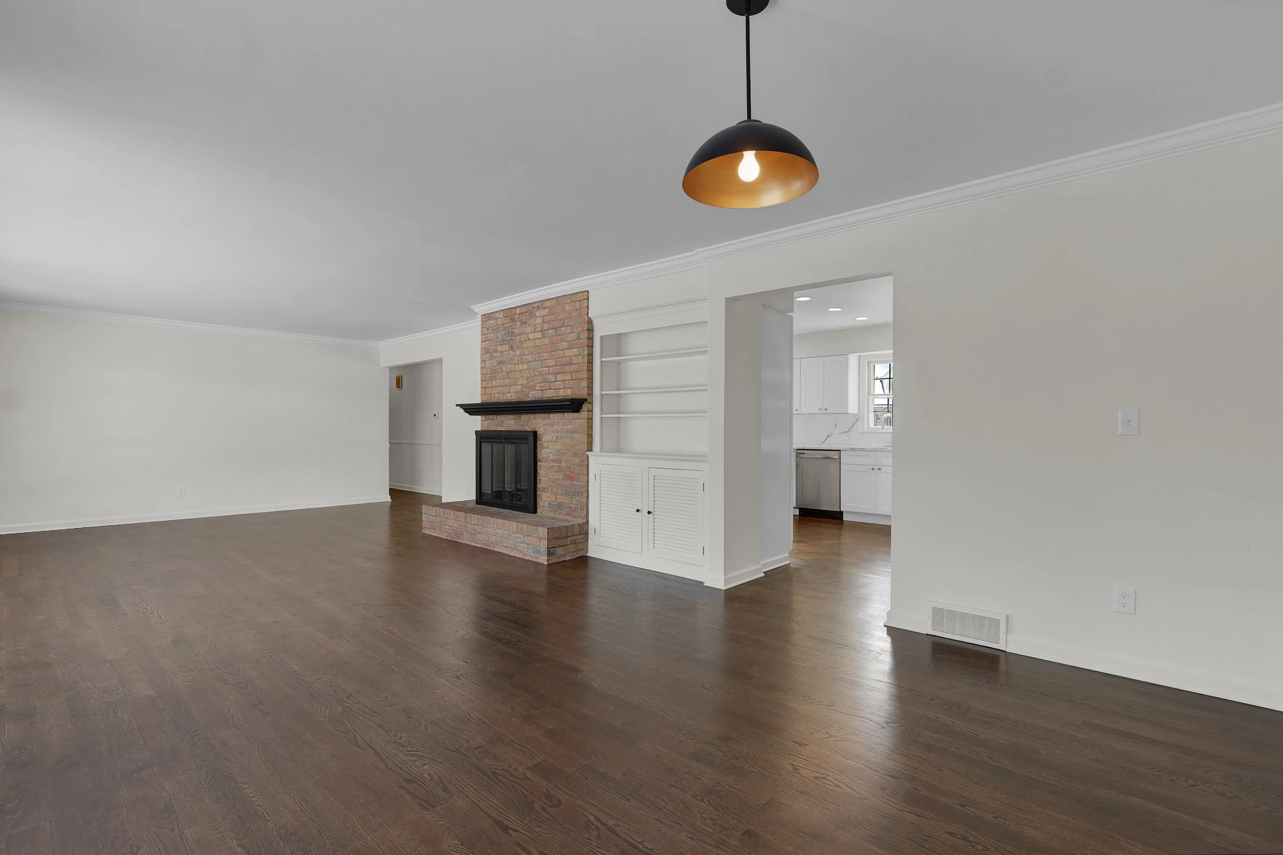 Empty living room with dark wood floors, white walls, a brick fireplace, and a black ceiling lamp.