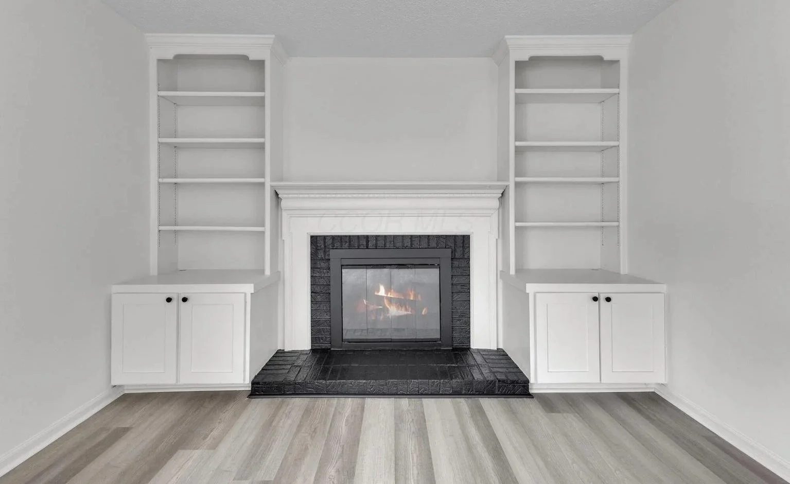Empty white built-in bookshelves around a black fireplace with a burning fire, in a minimalist living room with light wood flooring.