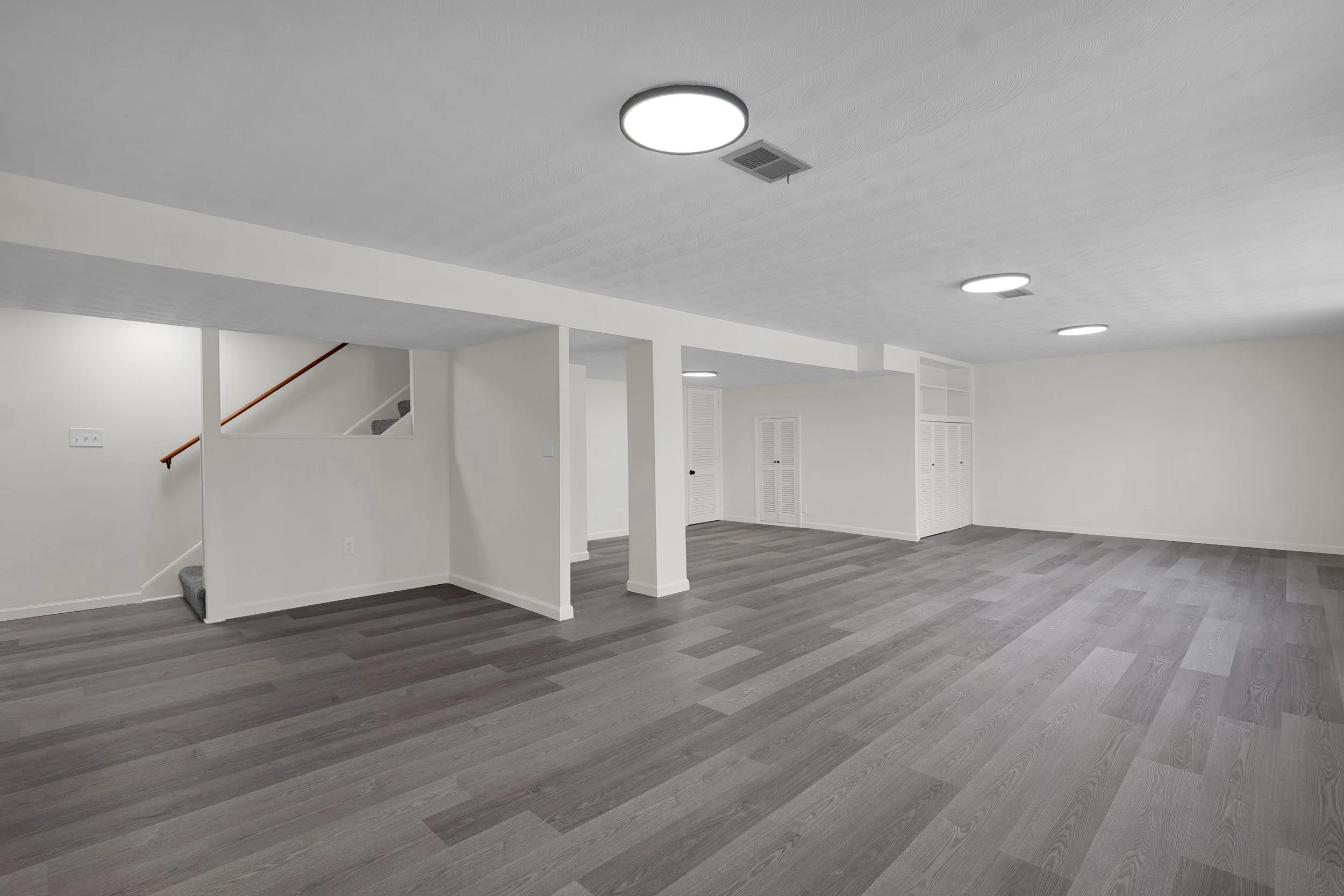 Empty living room with gray wood floors, white walls, a staircase with a wooden handrail, and ceiling lights.