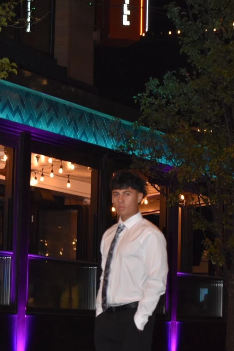 A young man in a white shirt and patterned tie standing in front of a building with colorful lighting at night.