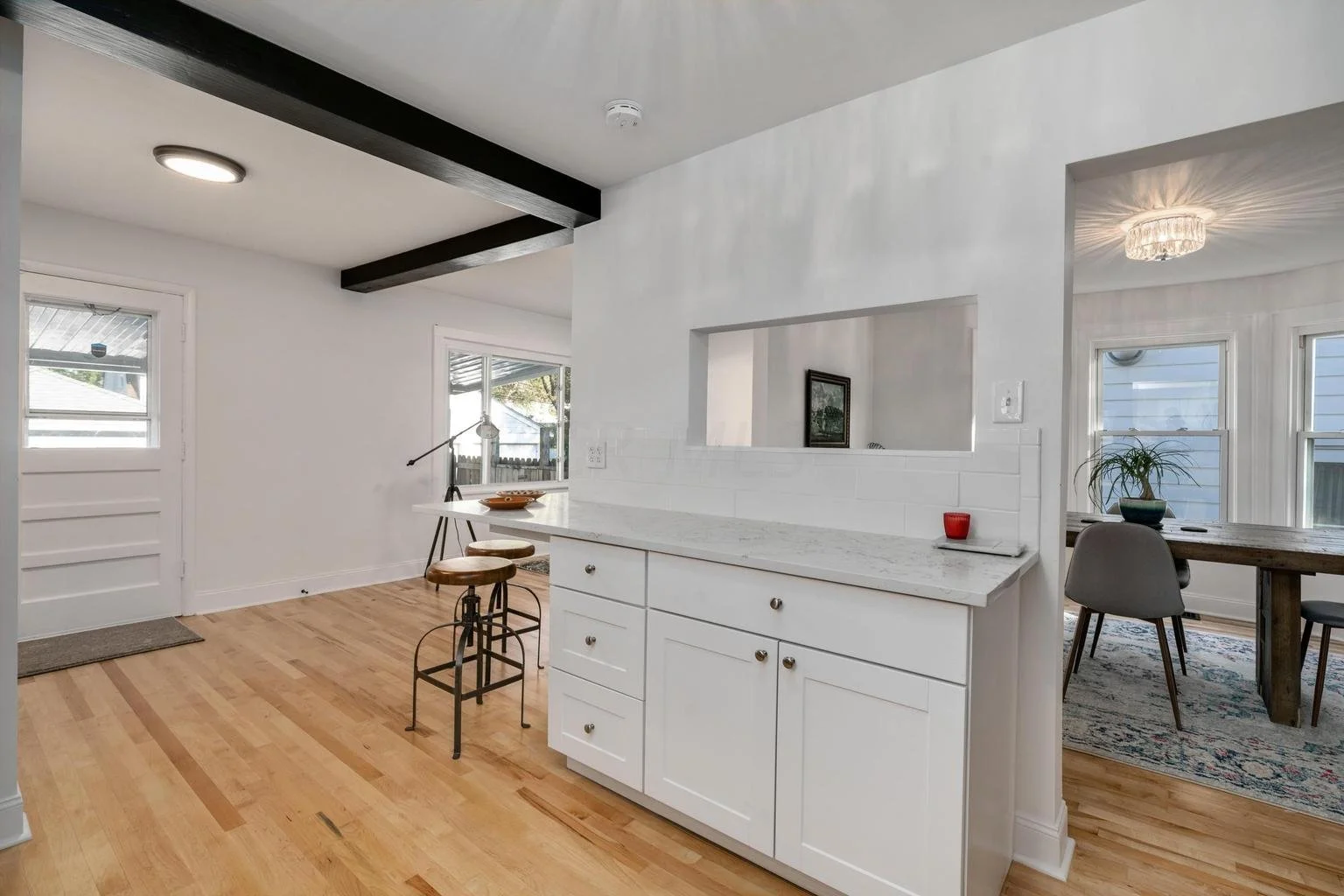 Open-concept kitchen and dining area with white cabinetry, wooden floors, and large windows, featuring a breakfast bar with three stools, a dining table with chairs, and decor such as a plant and a red candle.