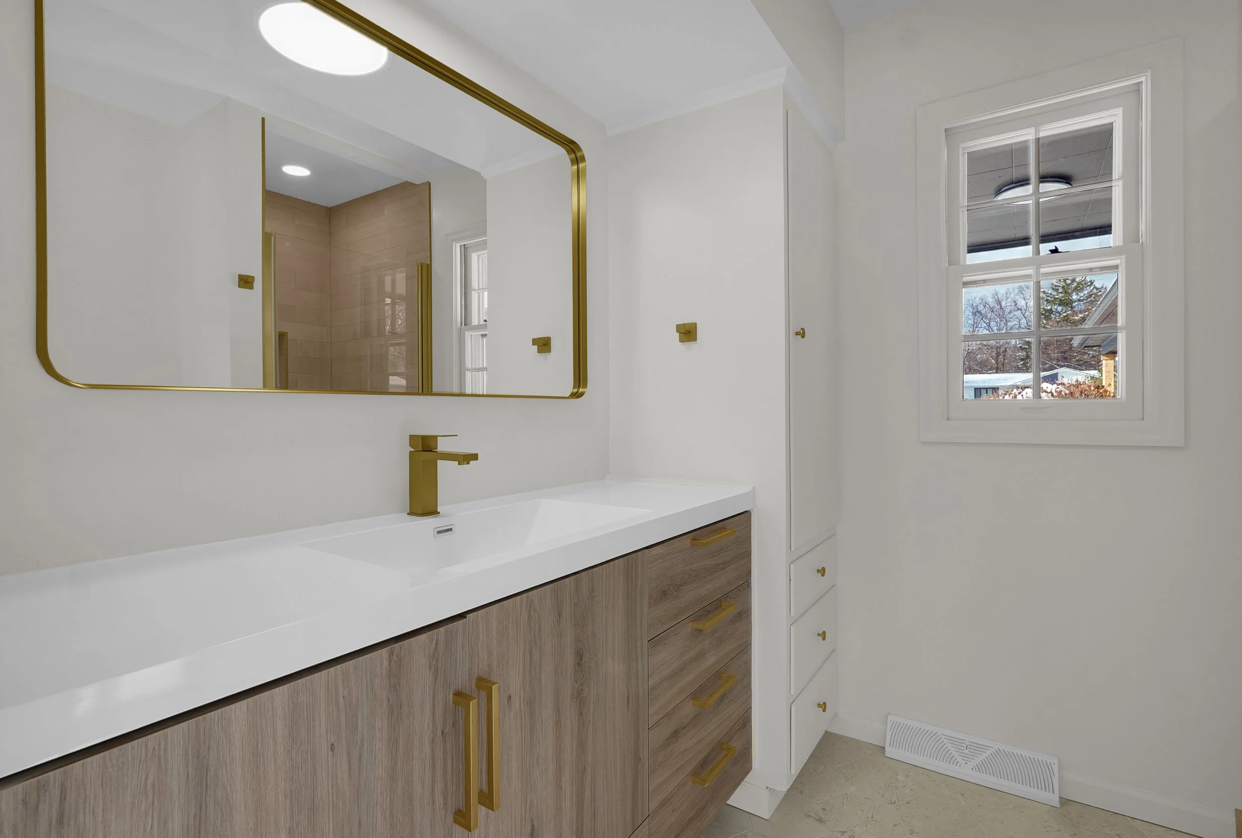 Modern bathroom vanity with a white countertop, wooden lower cabinets with gold handles, a large wall-mounted mirror with gold trim, a gold faucet, and a window showing a snowy outdoor scene.