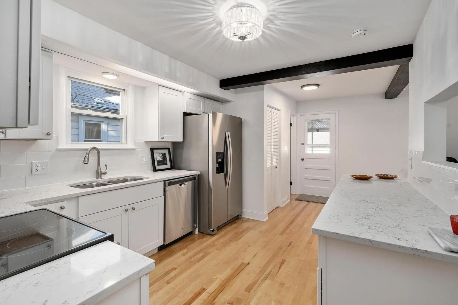 Bright modern kitchen with white cabinets, marble countertops, stainless steel refrigerator, and hardwood floor. A window above the sink, stylish lighting fixture, and black wooden beam on ceiling.