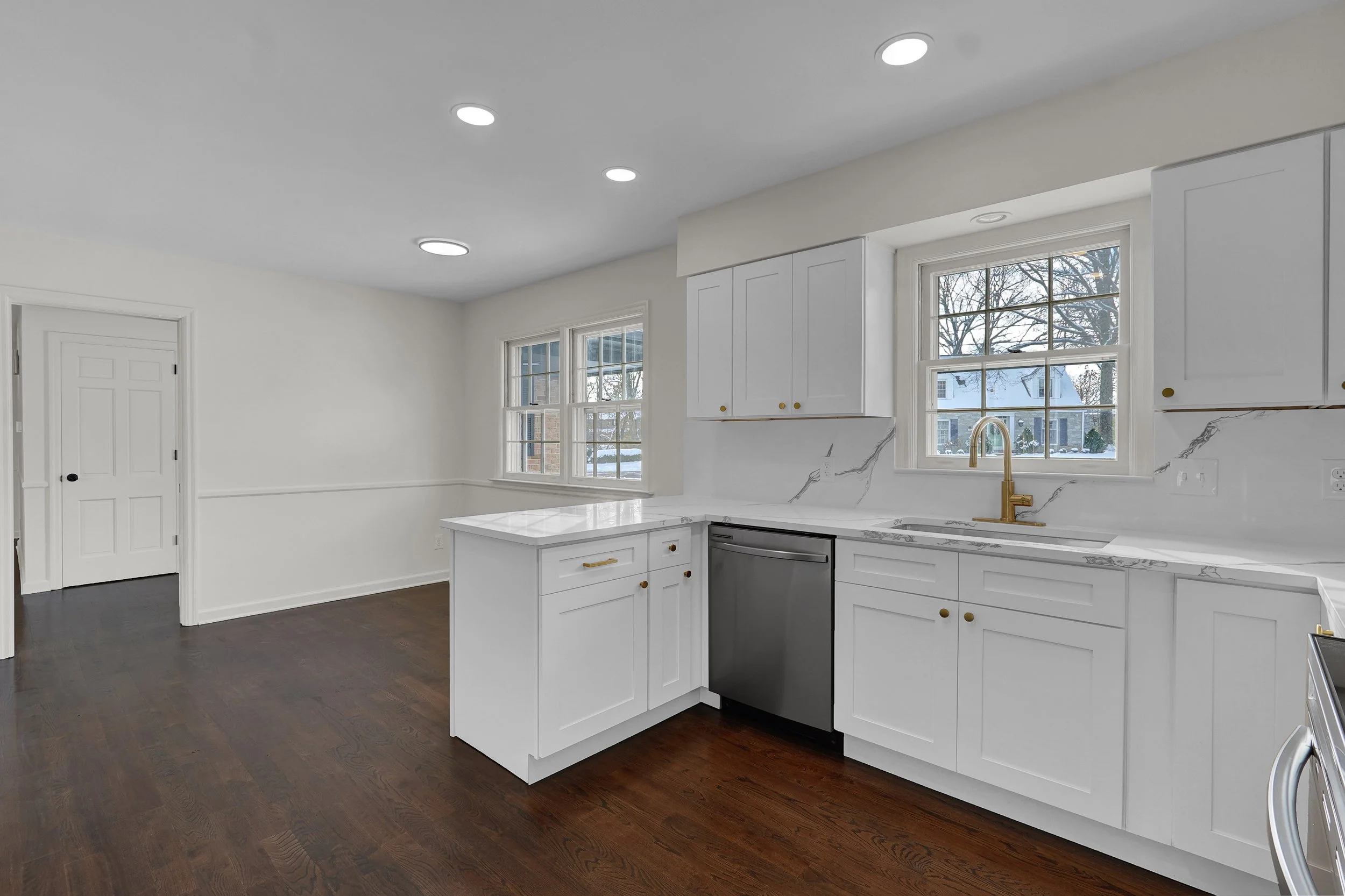 Modern white kitchen with marble countertops, gold hardware, dark hardwood floors, and natural light through large windows.
