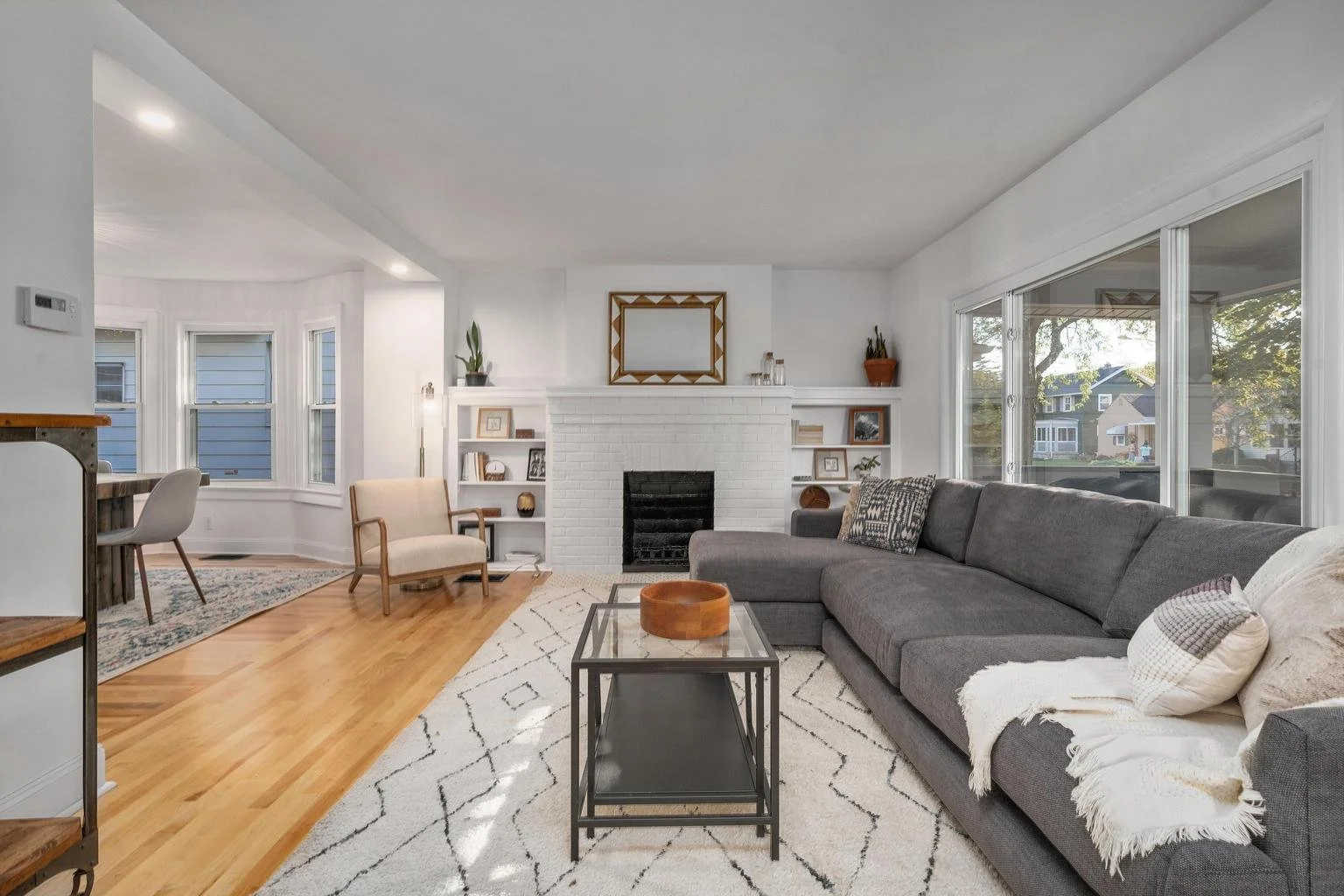 Living room with gray sectional sofa, black coffee table, white rug with black geometric patterns, white brick fireplace, large windows, and built-in shelves with decor.