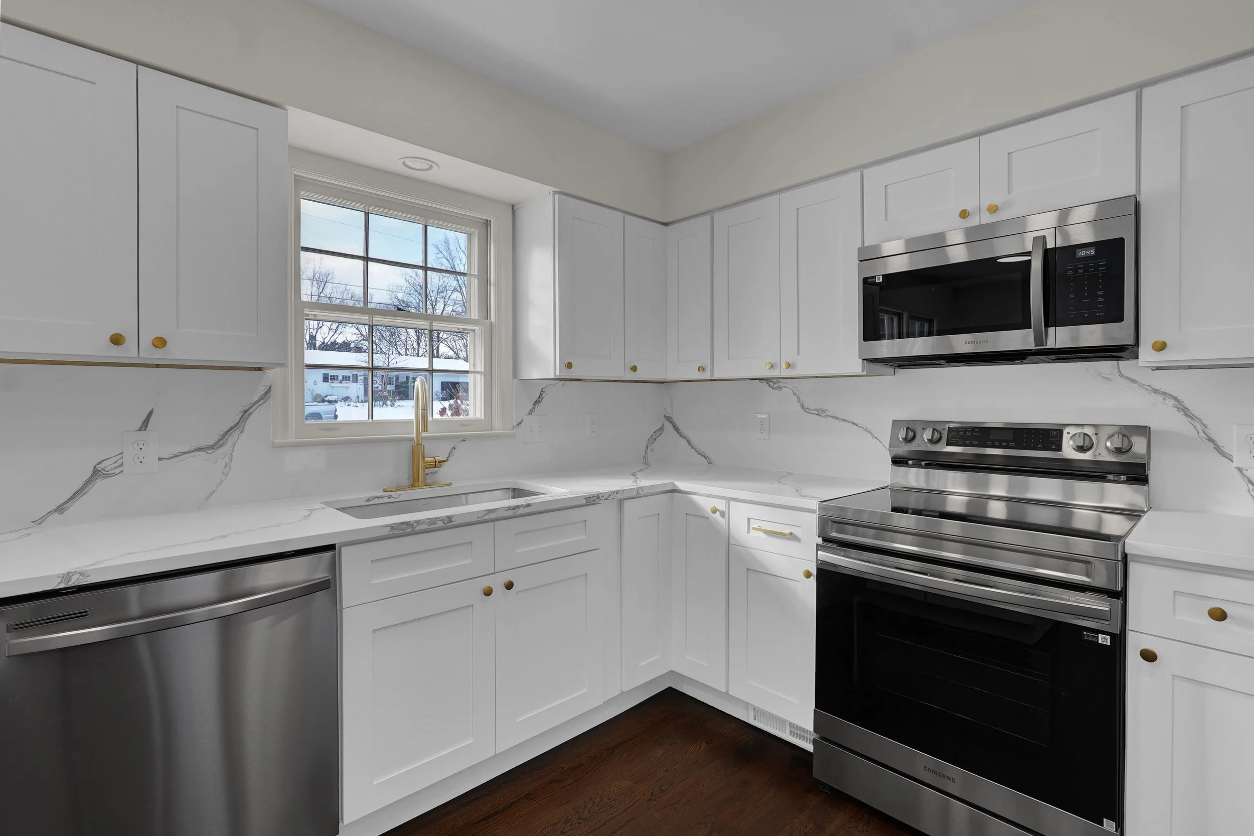 Modern kitchen with white cabinets, marble countertop and backsplash, stainless steel appliances including microwave and oven, gold fixtures, and a window showing snowy outdoor scene.