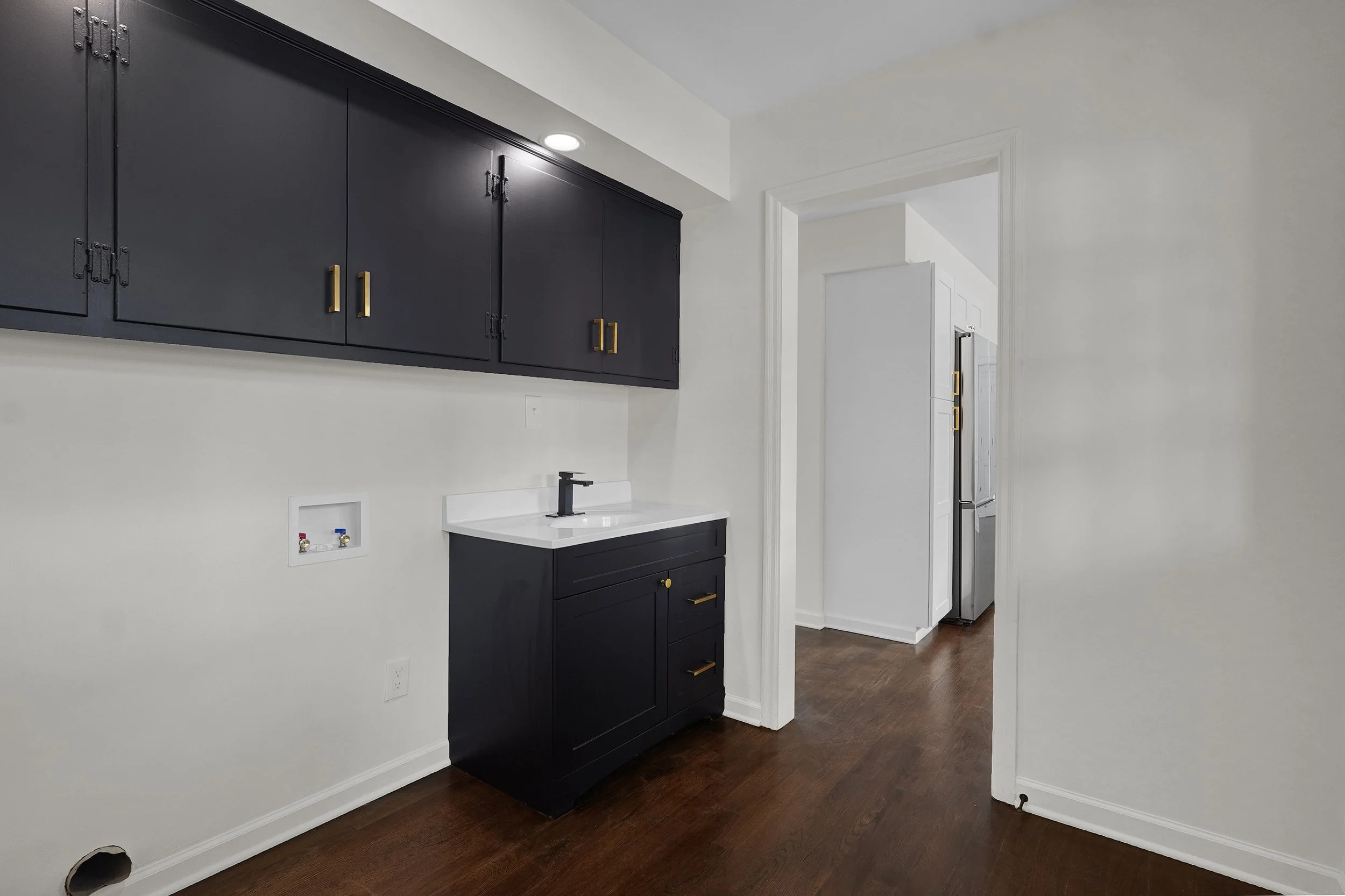 A kitchen with dark blue cabinets, a white countertop, and a black faucet. There is a washer or dryer hookup on the wall, and a doorway leads to another room with a fridge visible.