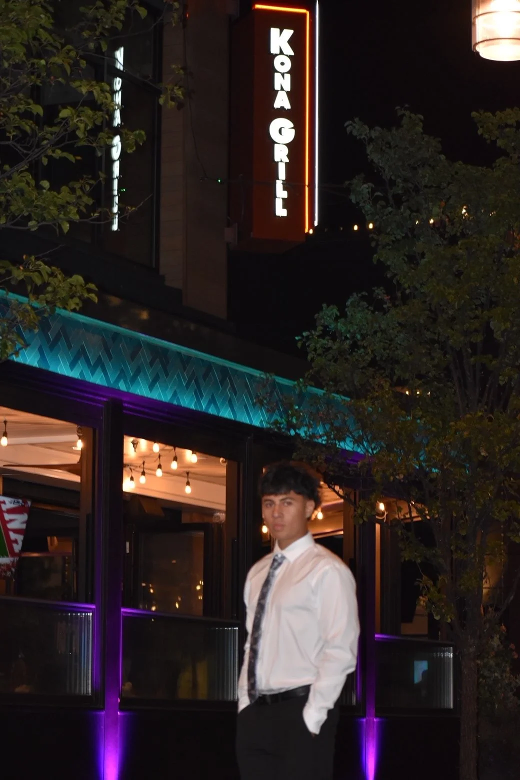 Young man in white shirt and patterned tie standing outdoors at night in front of a restaurant with neon sign that reads 'Kona Grill'.