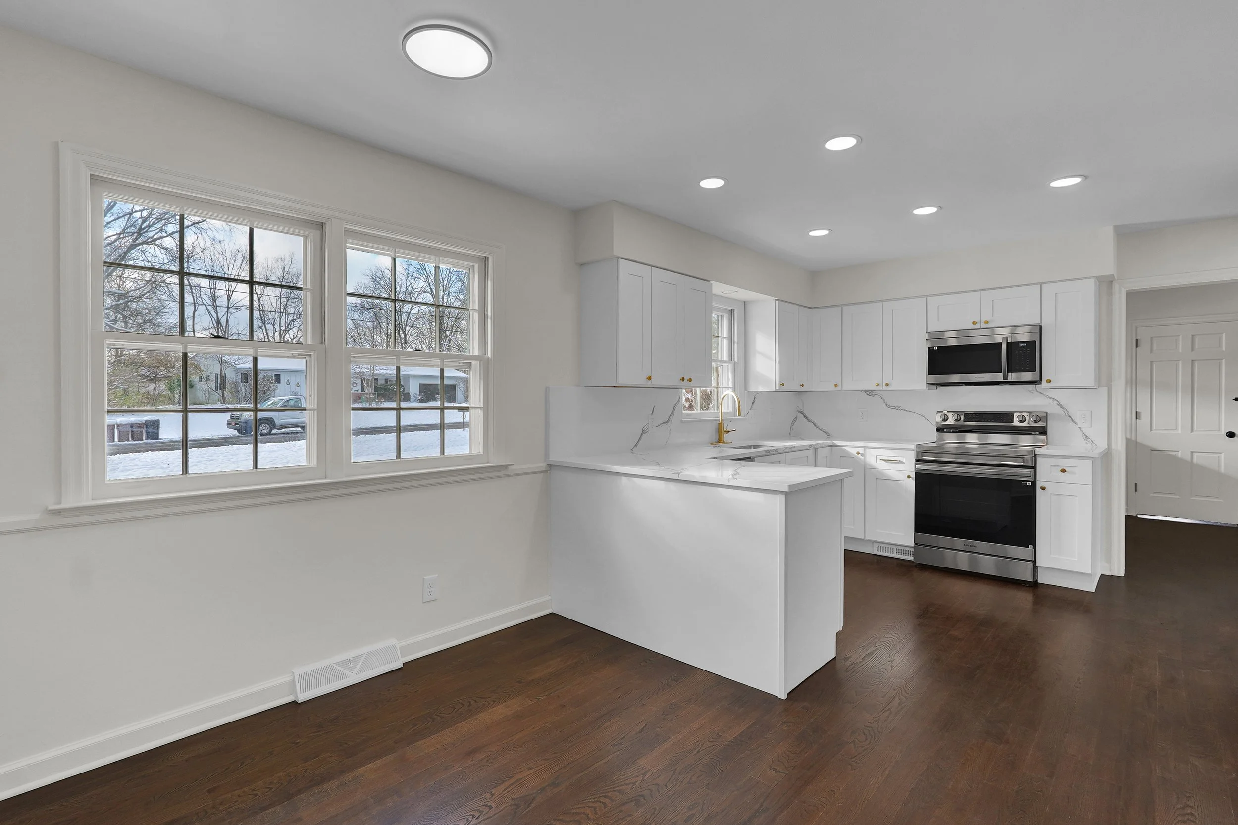 Interior of a modern kitchen with white cabinets, stainless steel oven and microwave, marble countertops, and large windows showing a snowy outdoor scene.