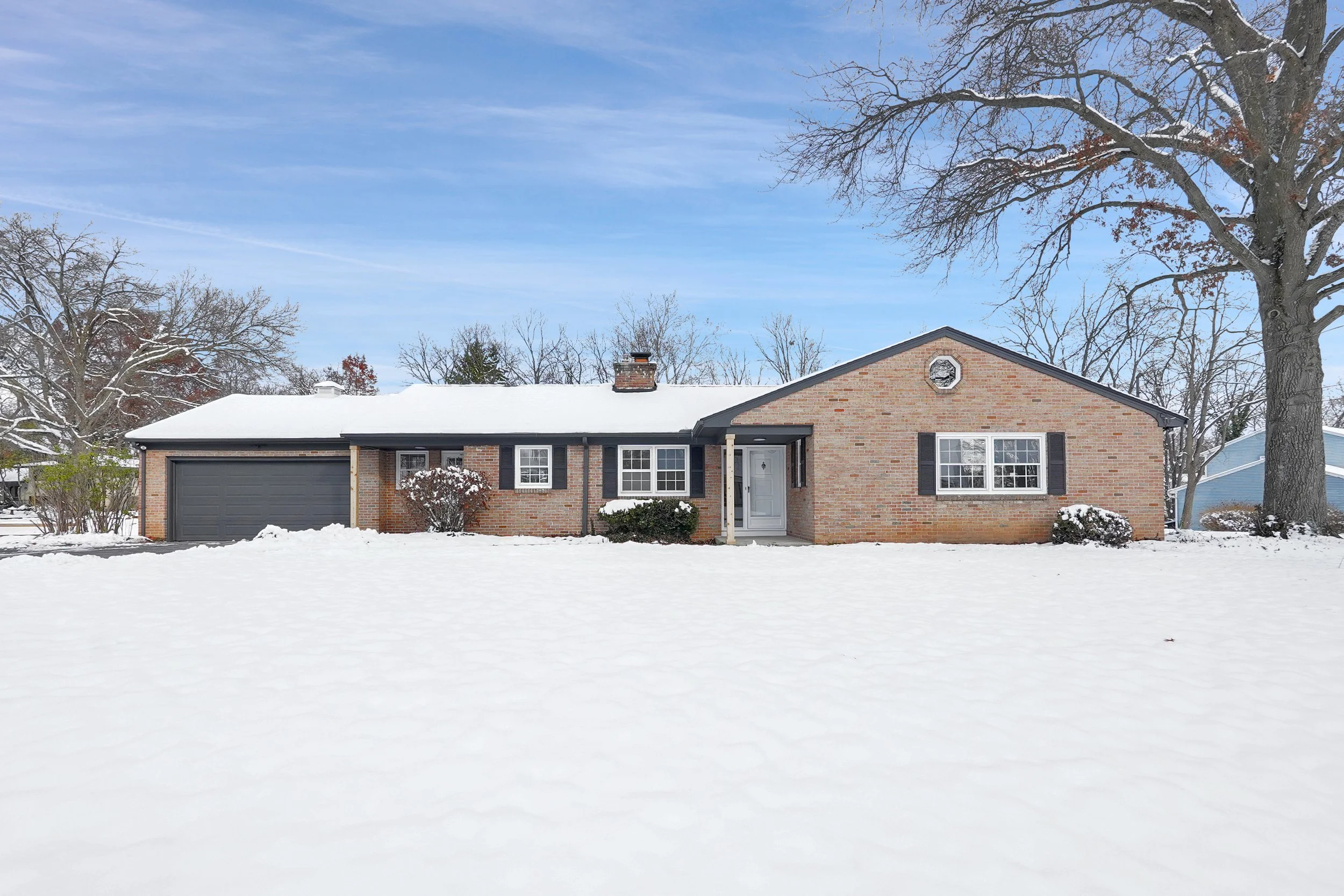 A single-story brick house with a snow-covered front yard and a large tree without leaves in winter. The house has black shutters, a white front door, a brick chimney, and an attached garage. The sky is partly cloudy with light, wispy clouds.