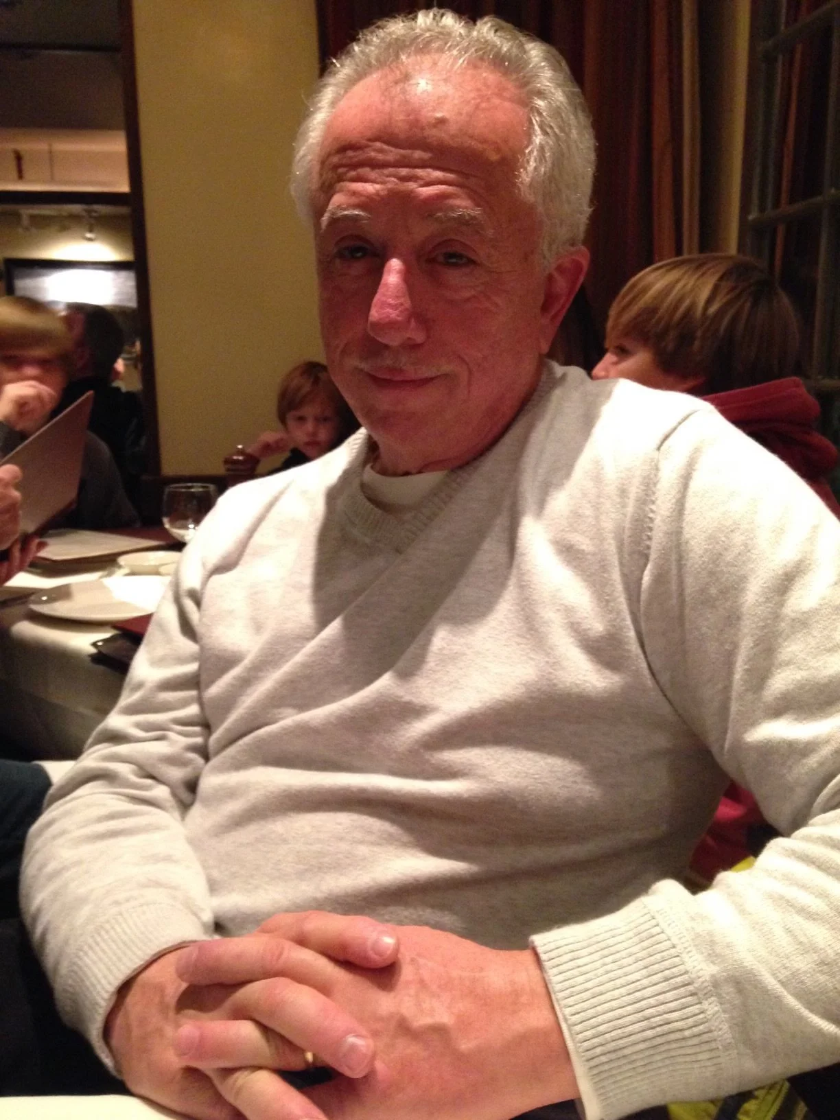 An older man with gray hair and a slight smile, wearing a light-colored sweater, sitting at a restaurant table with other people in the background.